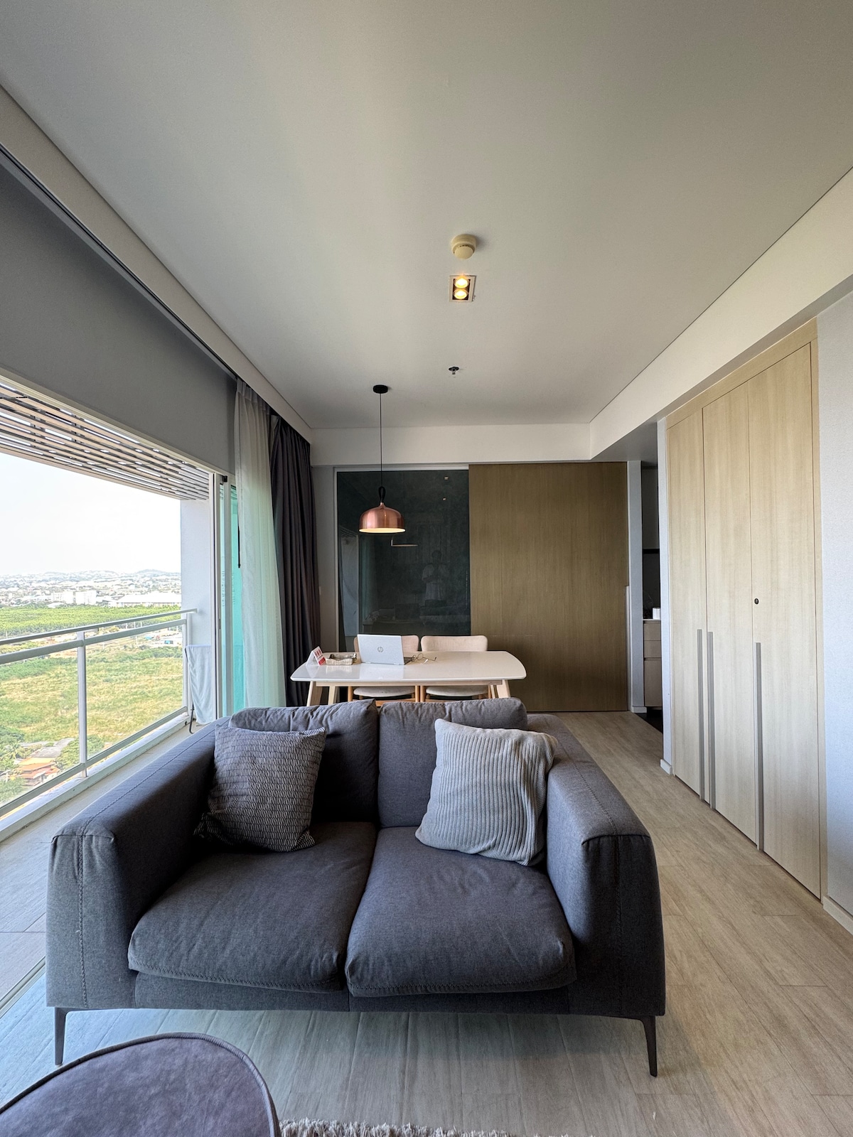 A modern living area features a comfortable gray sofa with decorative pillows, positioned by a large window offering views of the surrounding landscape. A minimalist dining table is visible in the background, accompanied by stylish lighting fixtures and sleek cabinetry.