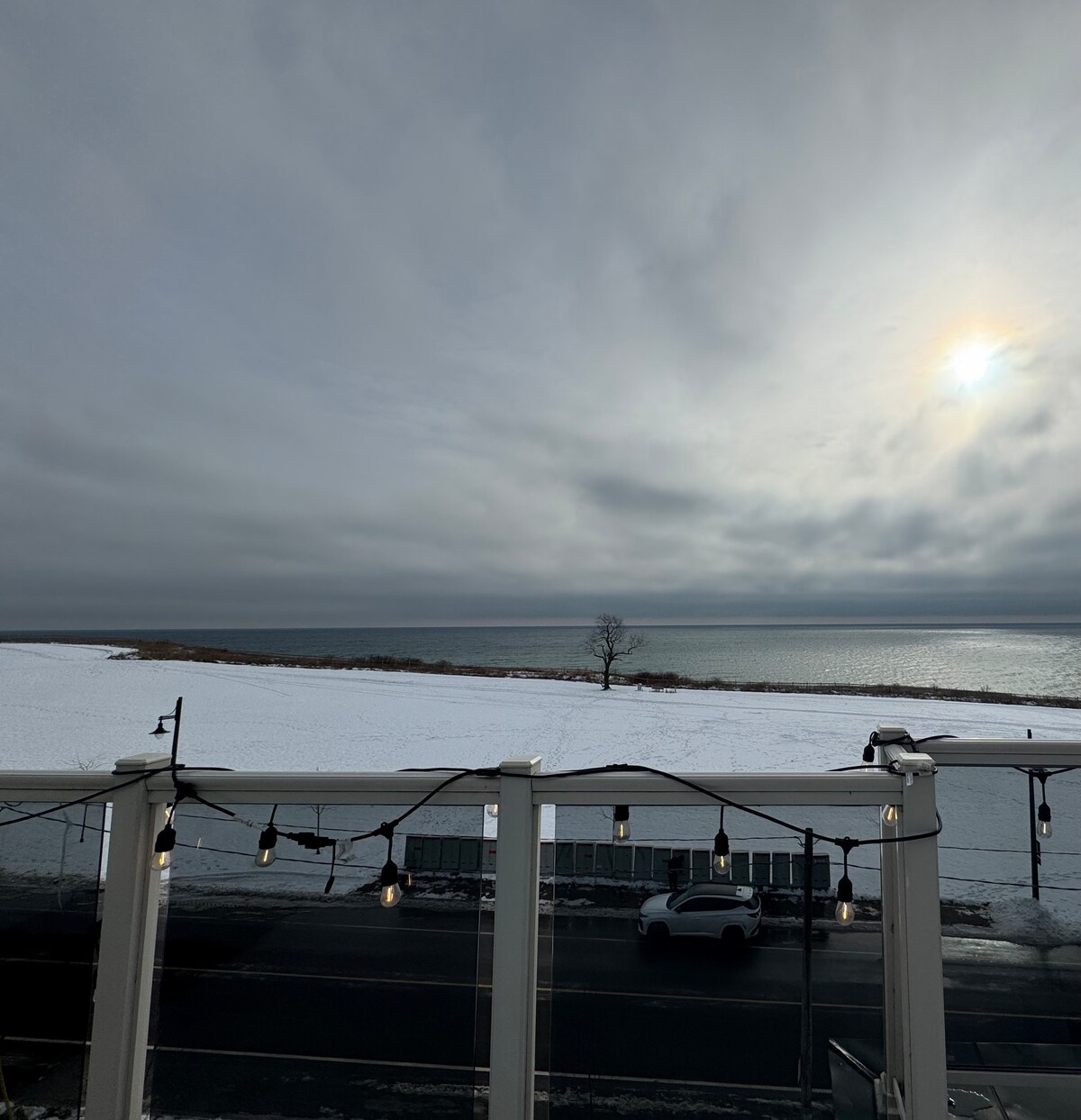 A serene view of the expansive Lake Ontario shoreline is visible, framed by a snowy landscape. The scene captures a cloudy sky with hints of sunlight peeking through. Softly glowing lights hang from a railing in the foreground, enhancing the tranquil atmosphere.