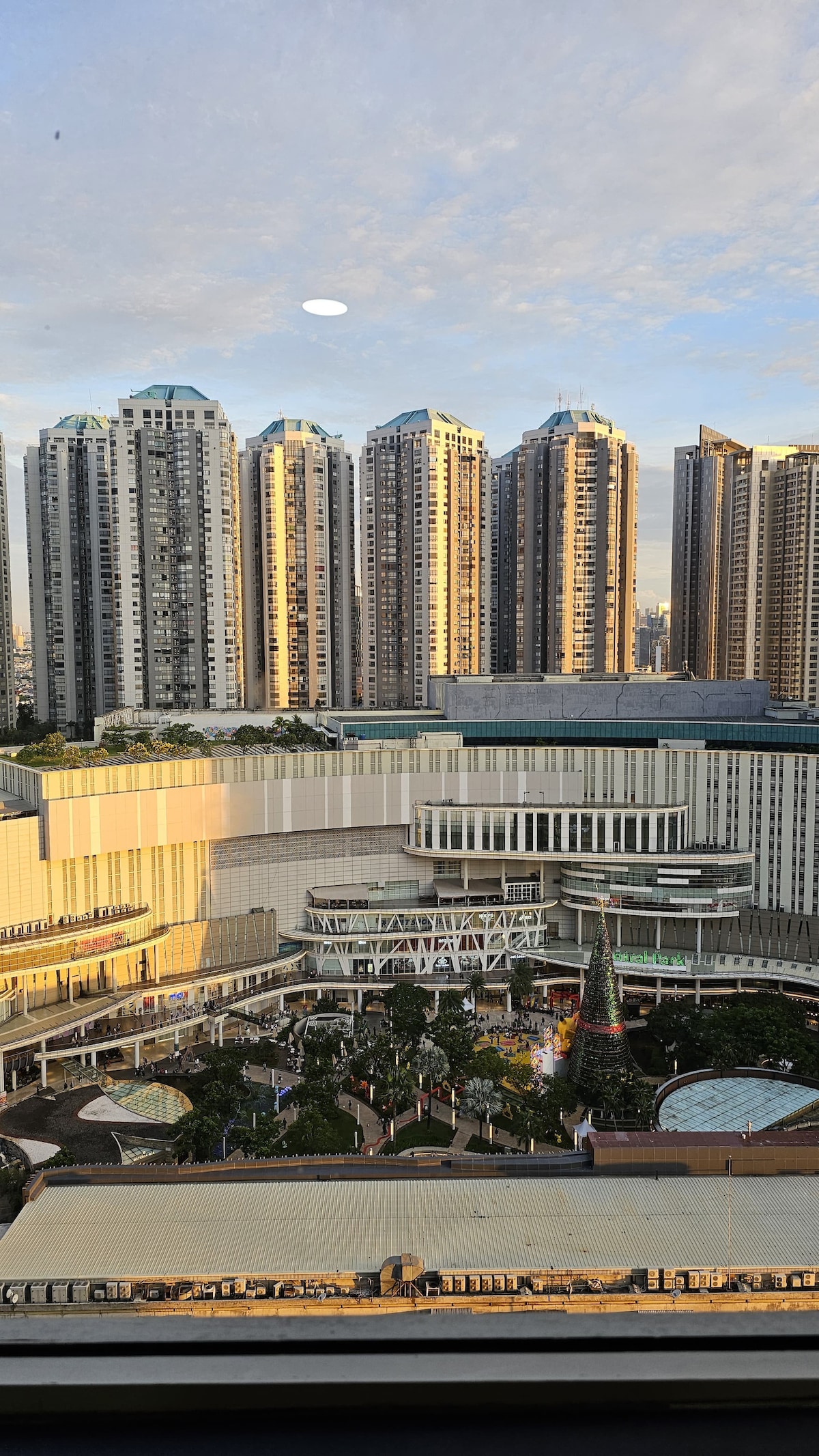 A panoramic view of the bustling Central Park Mall is displayed from the apartment balcony. Surrounding high-rise buildings reflect the golden light of the setting sun. Lush greenery and open spaces are visible in the mall's courtyard, highlighting a vibrant urban atmosphere.
