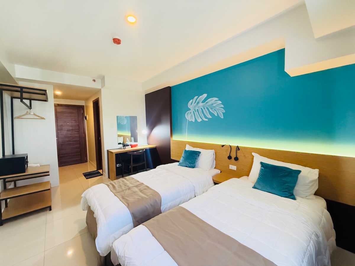 A well-lit studio unit features two spacious beds with light bedding, complemented by aqua-colored wall accents. A work desk and chair are present beside a mounted mirror. A minimal shelving unit adds storage, while a door leads to the entrance, enhancing accessibility.
