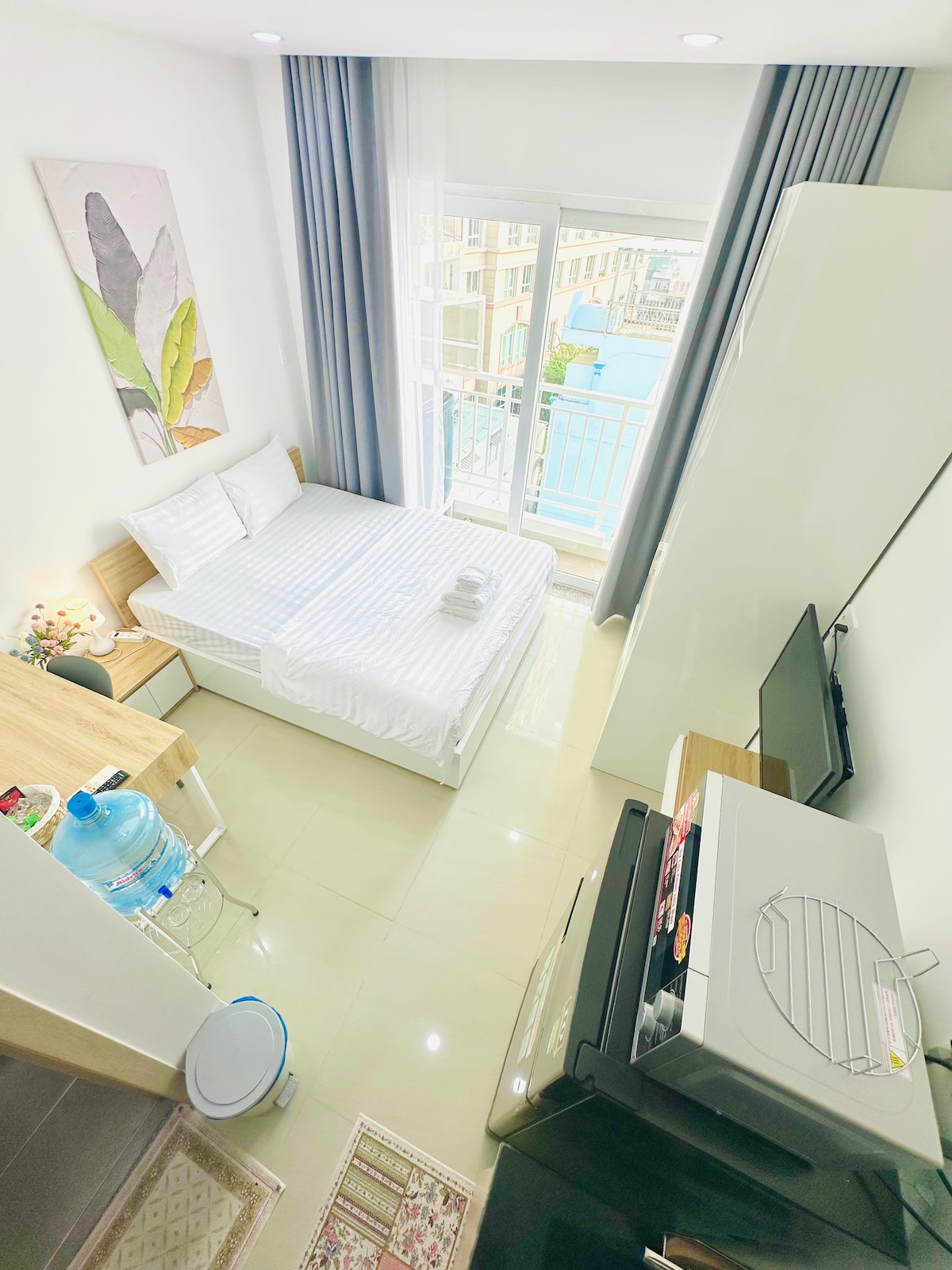 A bright and inviting sleeping area features a spacious bed with crisp white linens. A small work desk, a television, and a refrigerator are visible in the room. Large windows allow natural light to fill the space, providing a view of the outdoor surroundings.