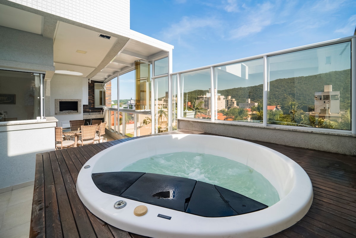 A private jacuzzi is positioned on a wooden deck, surrounded by glass walls that provide expansive views of the landscape. The serene water is visible, reflecting the sunlight, while an outdoor seating area is situated nearby, creating an inviting space for relaxation.