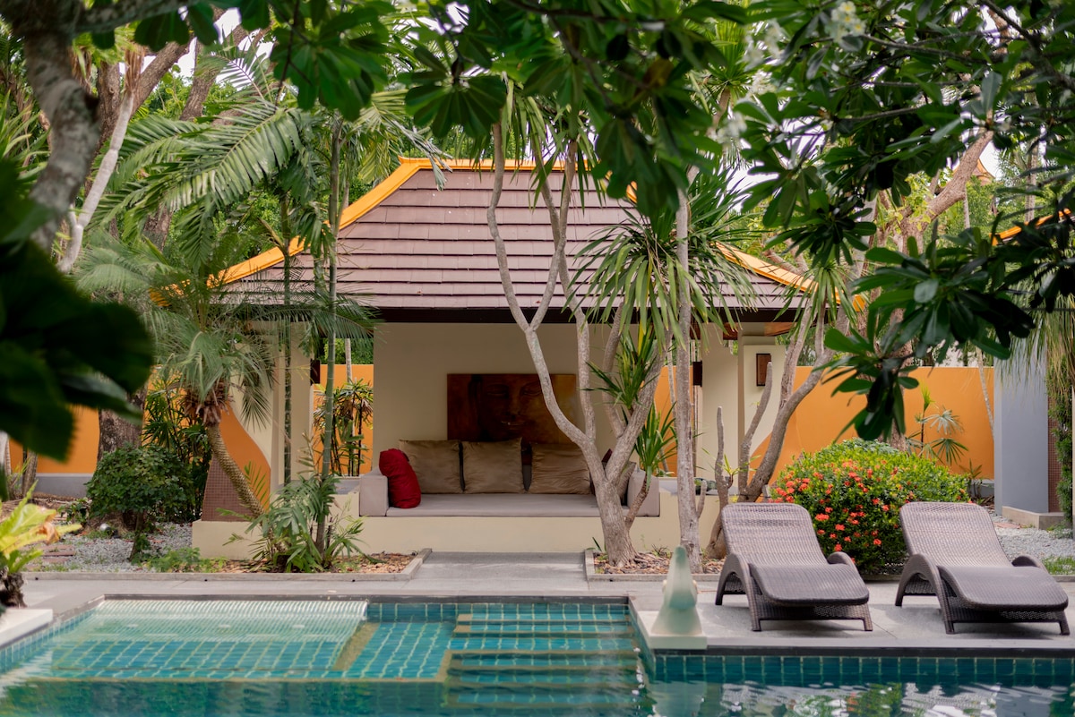 A private villa is framed by lush tropical greenery, showcasing an inviting outdoor seating area with soft cushions beneath a shaded porch. A tranquil swimming pool is visible in the foreground, complemented by lounging chairs positioned nearby, all contributing to a serene atmosphere.