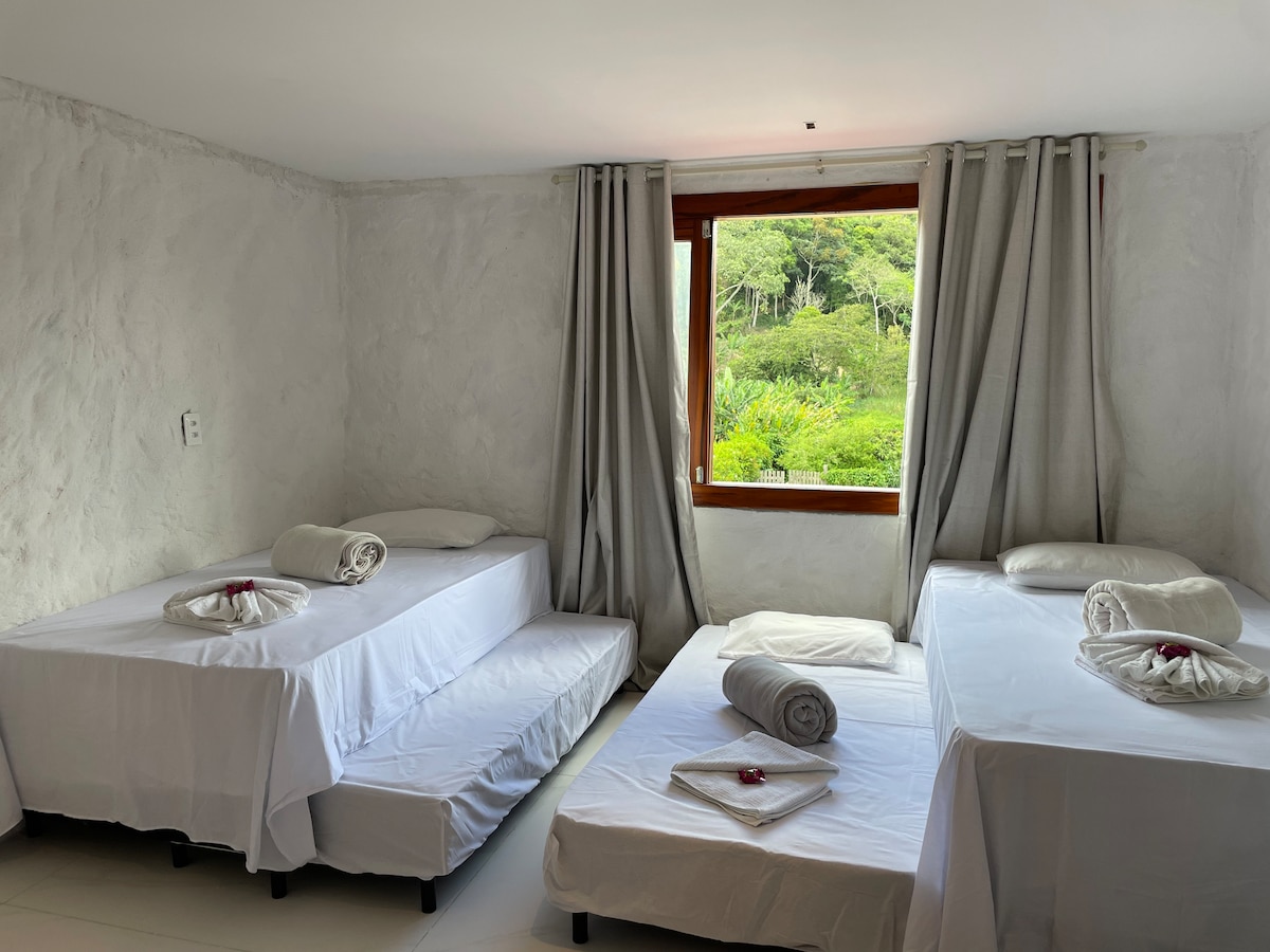 A light-filled room features a window overlooking a green landscape. Two single beds and one pull-out bed are neatly arranged, each dressed in white linens. Soft gray curtains frame the window, complementing the room's neutral walls.