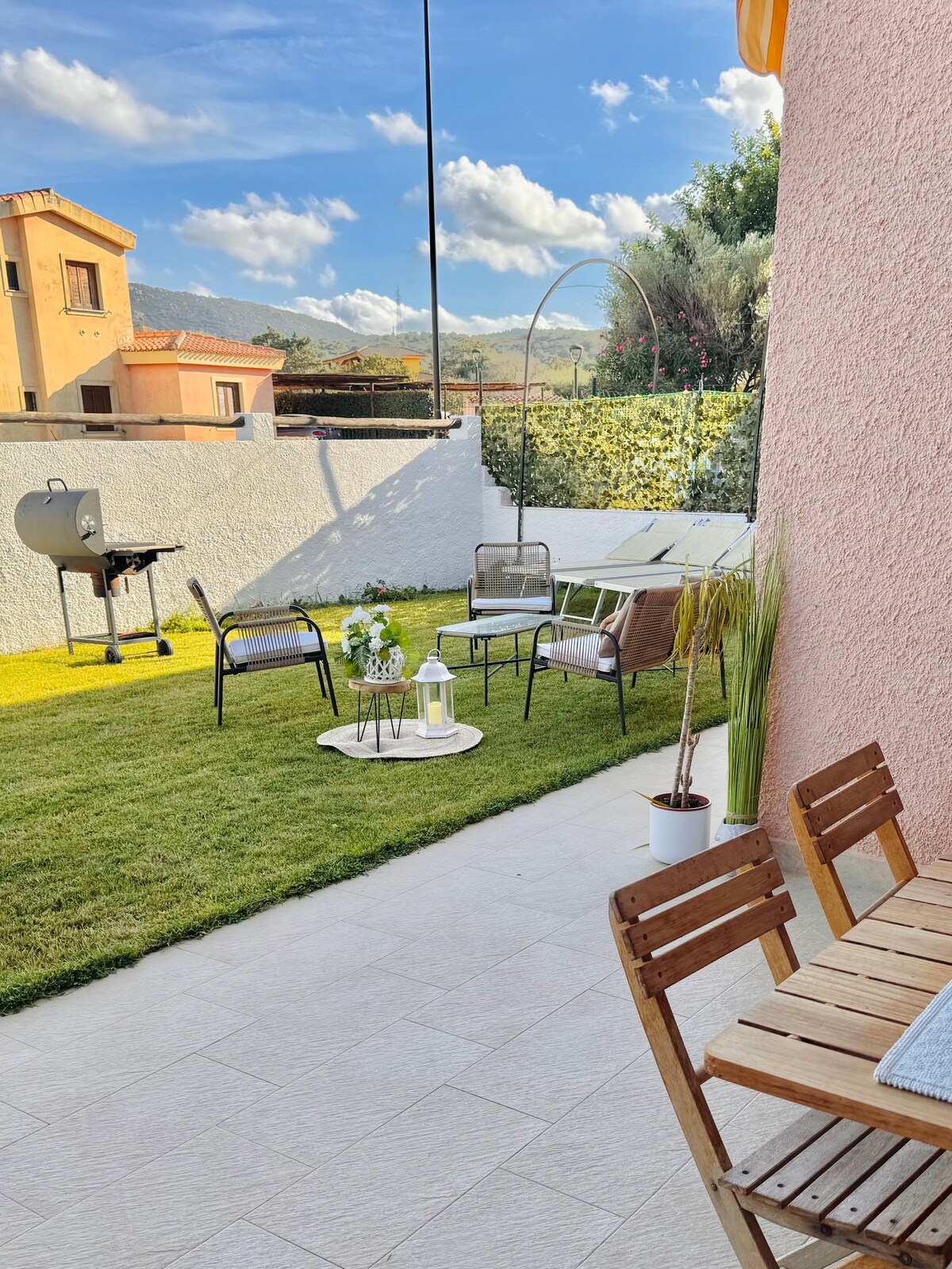 An inviting garden space features a green lawn with a barbecue grill in the background. Comfortable seating arrangements include two chairs and a coffee table surrounded by plants. A terrace area is visible, providing a relaxing outdoor atmosphere.