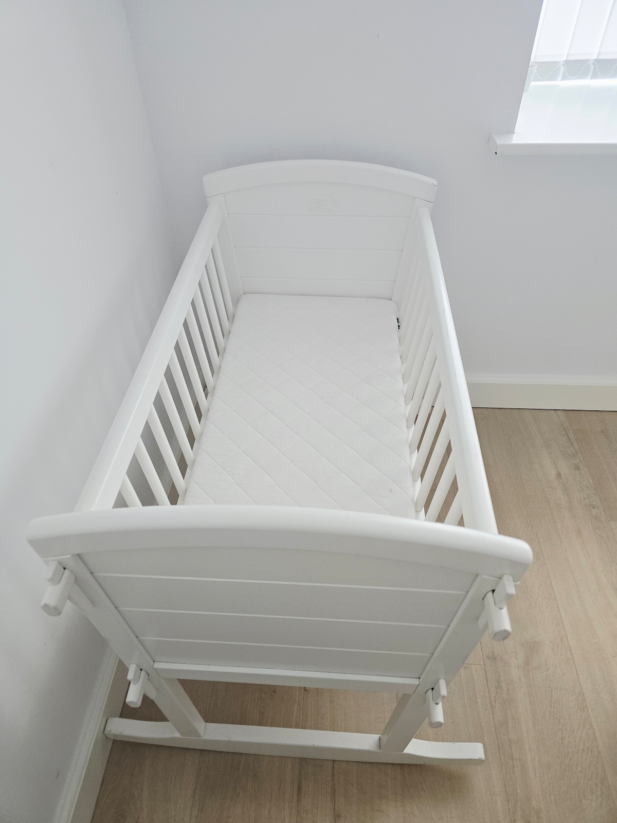 A simple white crib is displayed against a neutral wall, featuring slatted sides and a smooth, padded mattress. The room is illuminated by soft natural light entering through a nearby window, creating a calm and organized space.