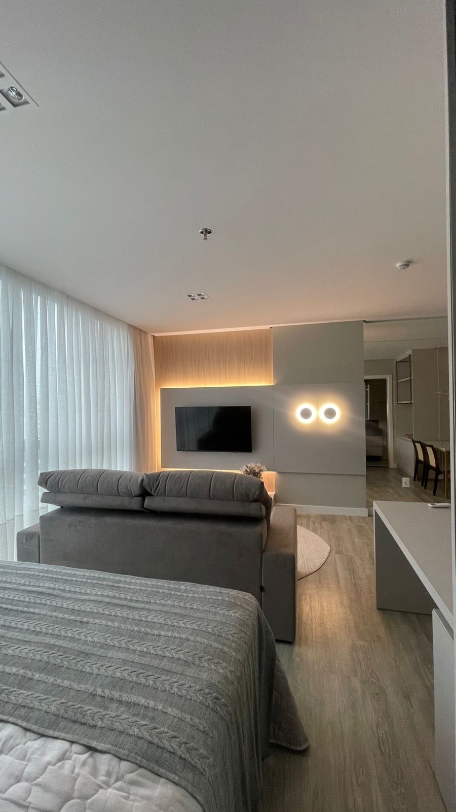 A modern living space features a plush gray sofa positioned against a light gray wall, accompanied by a mounted flat-screen TV. Natural light filters through sheer curtains, creating a soft ambiance. A sleek white desk is visible nearby, contributing to the room's contemporary design.
