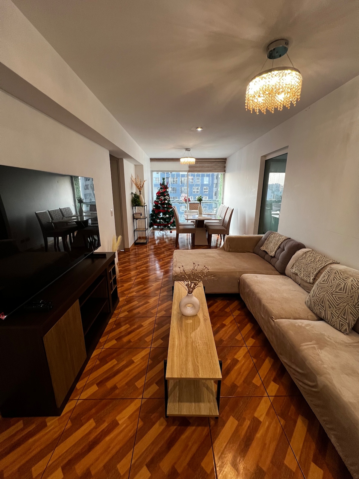 A living area showcases a sectional sofa paired with a minimalist coffee table. A television unit sits against a wall, while a dining area with a table and chairs is visible through large windows. A decorative Christmas tree adds a festive touch.