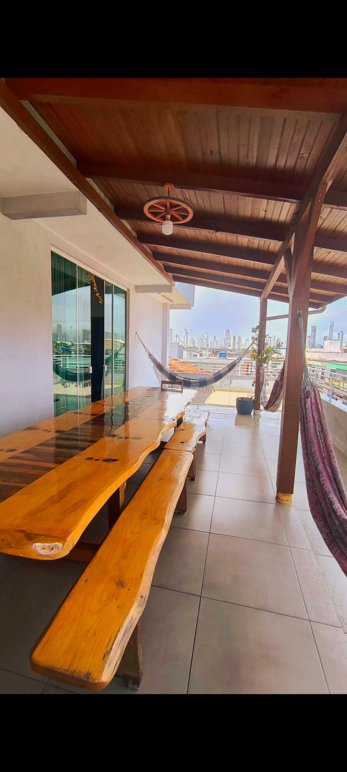 A spacious balcony area features a long wooden table with attached benches. Two hammocks are positioned for relaxation, with a backdrop of city views. Large glass doors provide access to the indoor space, while overhead beams create a welcoming atmosphere.