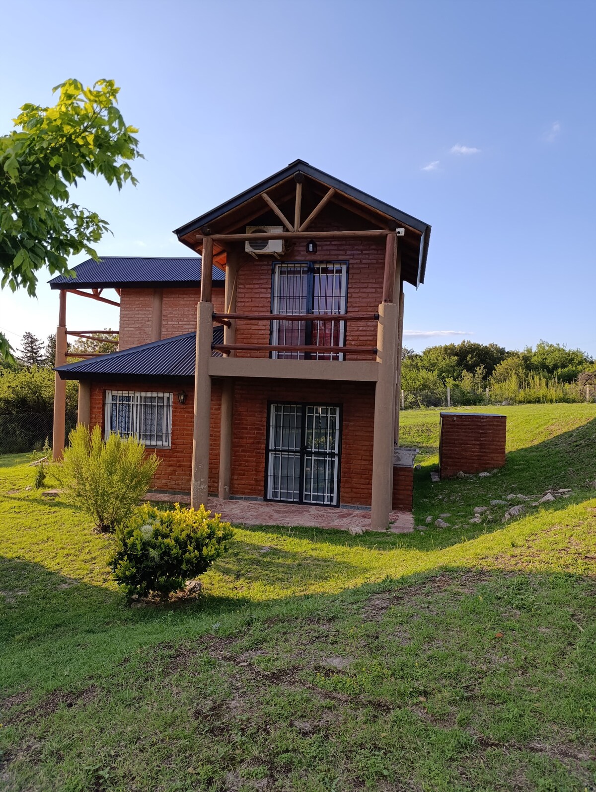 A two-story cabin is nestled amid lush greenery, showcasing a wooden exterior and a sloped roof. A balcony extends from the upper floor, offering views of the surrounding landscape. The well-maintained lawn features a variety of plants, enhancing the tranquil setting.
