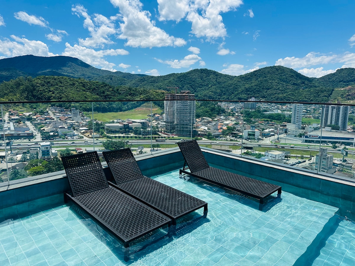An outdoor area features a sleek infinity pool with clear blue water. Two black lounge chairs are positioned on the pool's edge, surrounded by a glass railing that provides unobstructed views of the surrounding mountains and town below. Bright, sunny weather enhances the serene atmosphere.