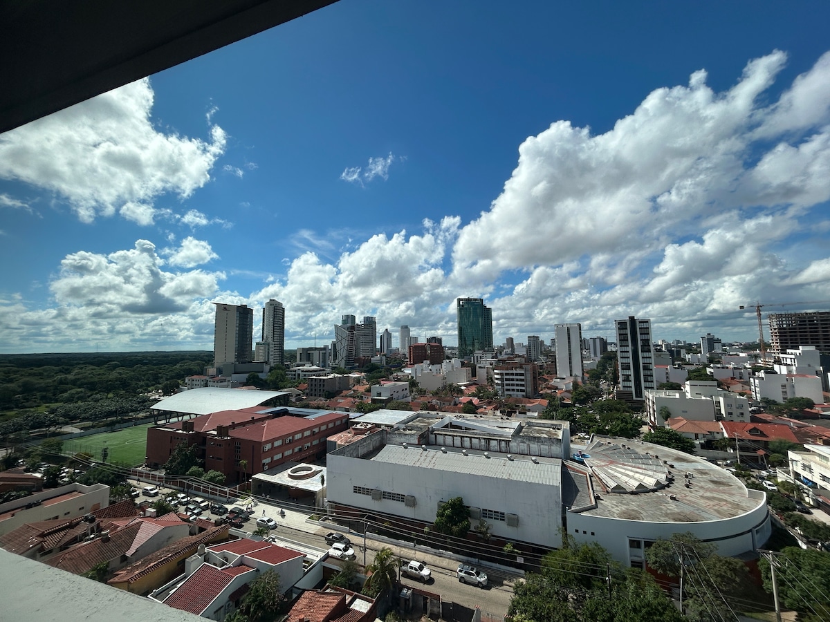 A panoramic view captures the city skyline, with modern high-rise buildings dotting the horizon under a partly cloudy sky. Below, urban structures and green spaces are visible, reflecting the vibrant atmosphere of the area.