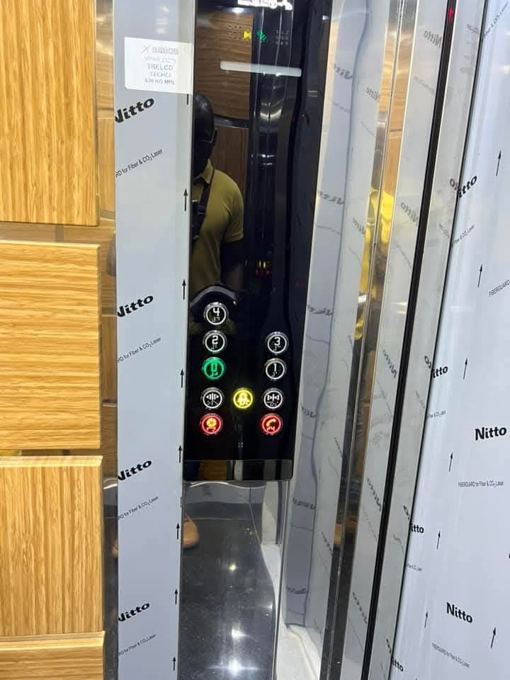 An elevator panel is displayed with illuminated buttons indicating various floor levels. The sleek design includes a reflective surface and wooden accents, contributing to a modern and elegant ambiance. Clear symbols assist with navigation, providing accessibility for all guests.