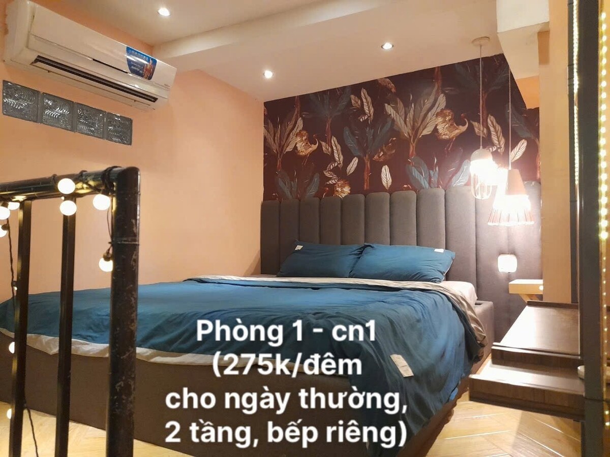 A comfortable bedroom features a spacious bed dressed in soft blue linens. The wall is adorned with a floral design. Soft lighting is provided by a pendant lamp, and an air conditioning unit is mounted above. The wooden floor adds a warm touch to the space.