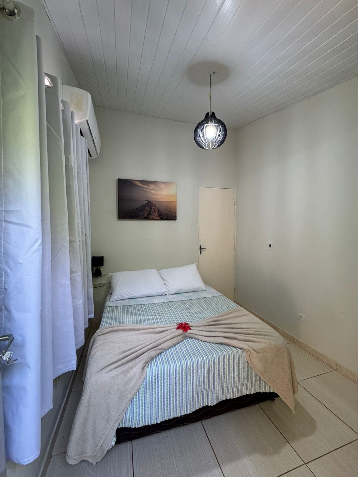 A cozy sleeping area features a well-made bed adorned with a soft blanket, positioned near a light-colored wall. A framed photograph hangs above the bed, and natural light enters through the window draped with sheer curtains, creating a tranquil atmosphere.