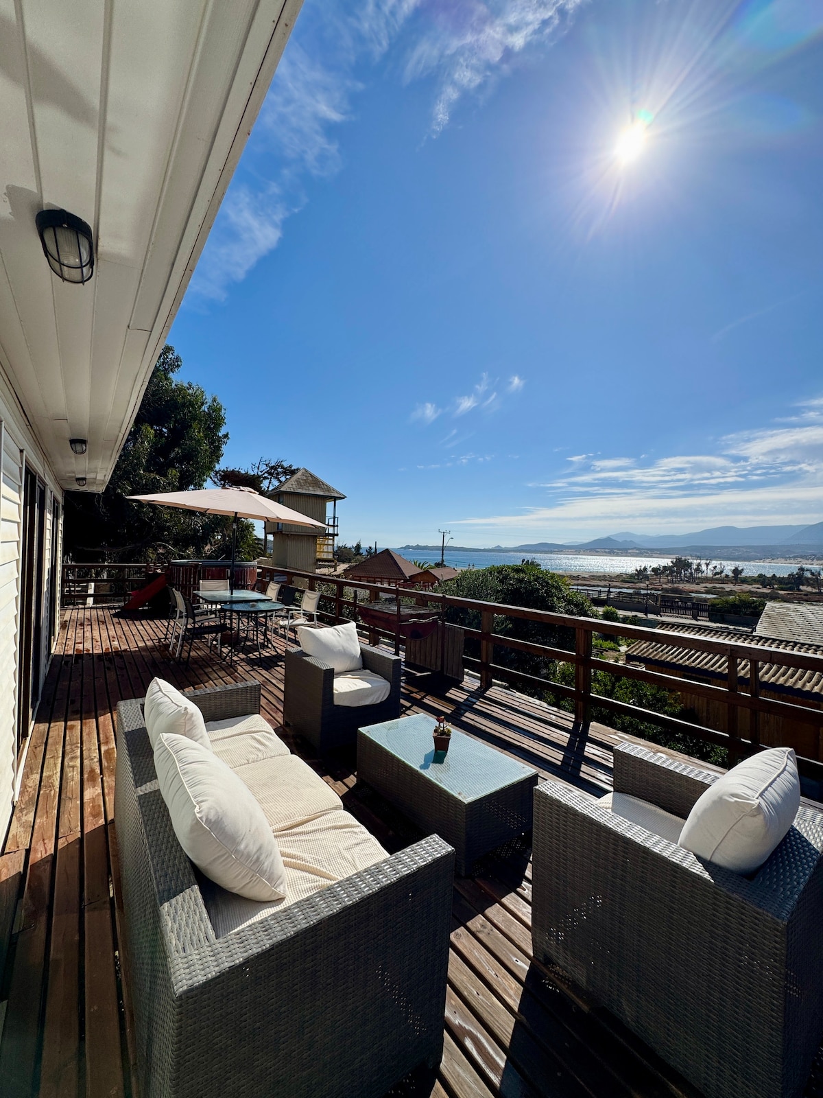 A spacious outdoor deck features comfortable seating arrangements with woven chairs and a glass-topped table. An umbrella provides shade, while the view includes distant hills and a glimpse of the water under a clear blue sky.
