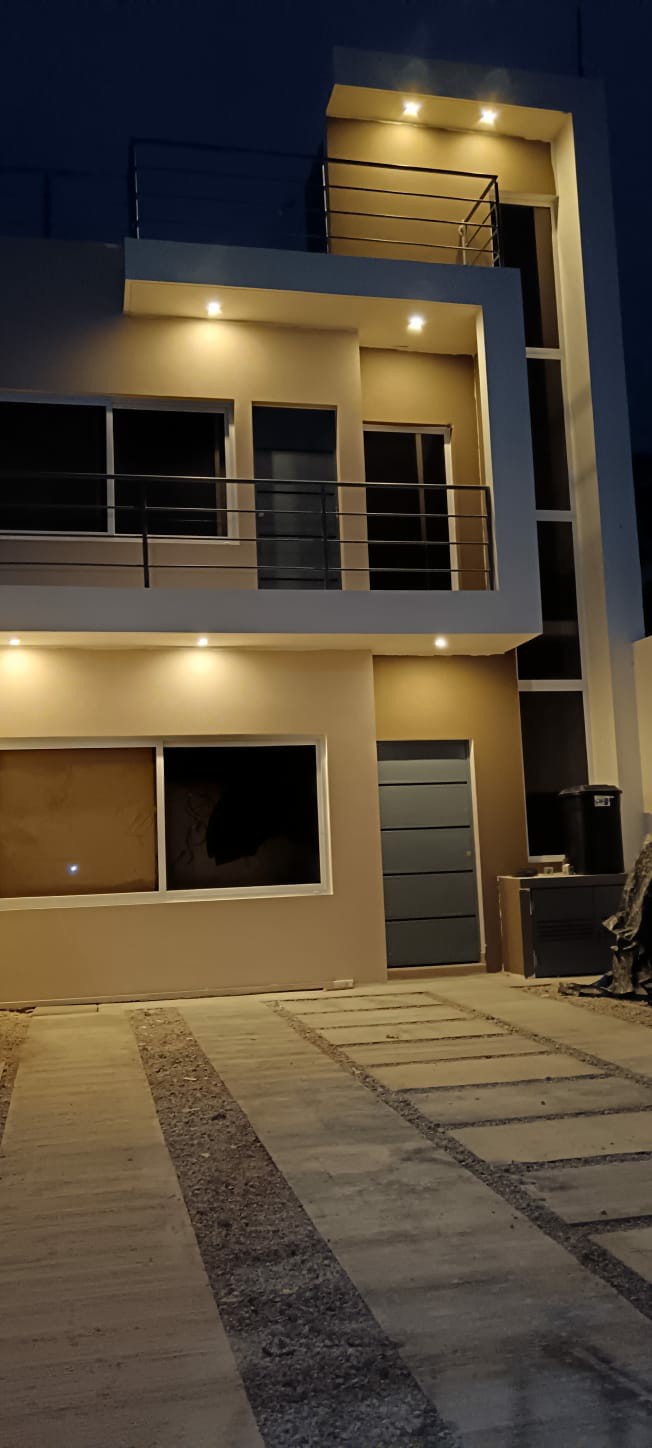 The modern two-story structure is illuminated at night, showcasing a clean façade with large windows and a door. A minimalist balcony extends from the upper level, while a paved area leads to the entrance.