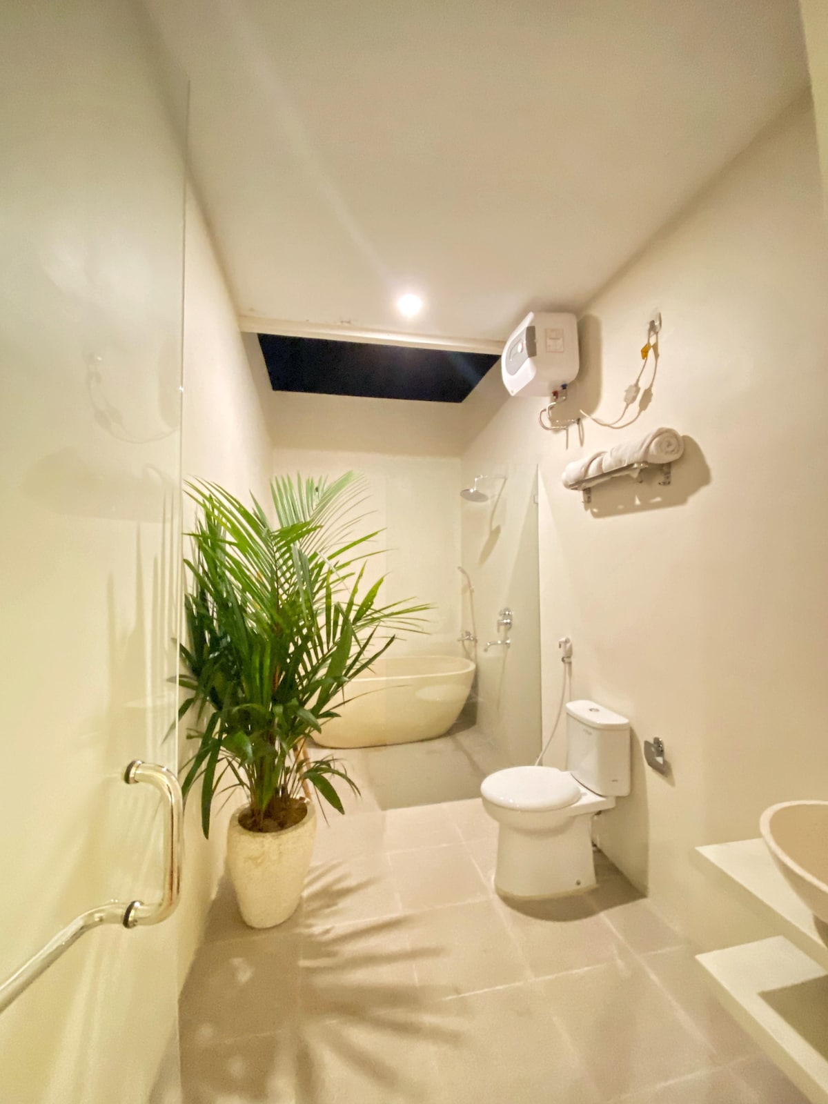 The bathroom features a spacious layout with a large bathtub and a separate shower area. A modern toilet sits adjacent to a decorative plant in a pot. Natural light filters in from the overhead space, enhancing the neutral color palette of the walls.