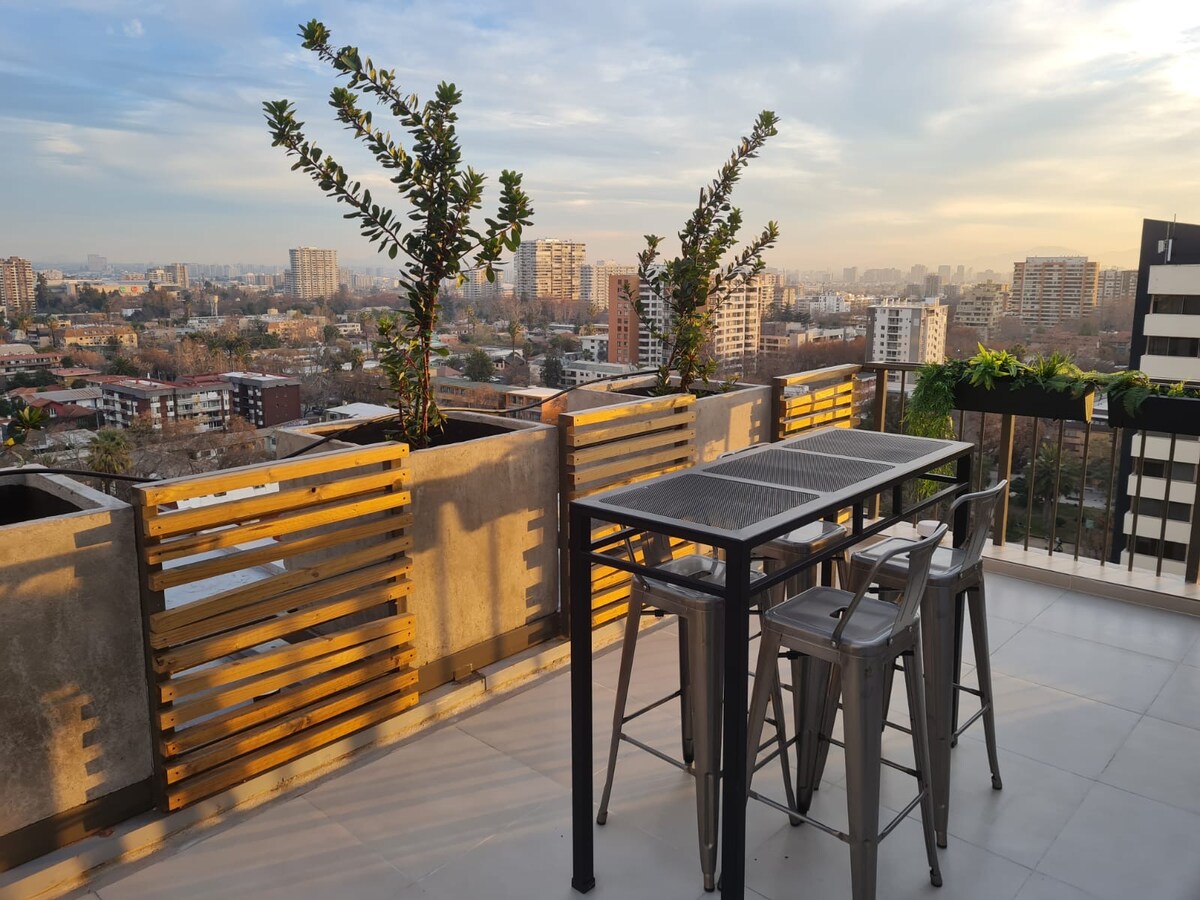 A rooftop terrace is presented, featuring a modern outdoor dining set with a table and four sleek chairs. Planters with greenery are positioned along the railing, providing a natural touch. The city skyline is visible in the background under a soft, evening sky.