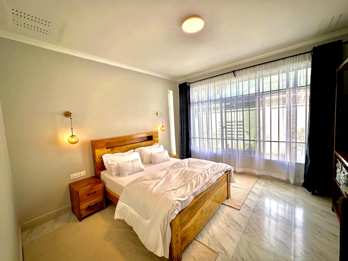 A spacious bedroom features a large wooden bed with crisp white bedding. Nightstands are positioned on either side of the bed, each with a light fixture. Natural light filters through large windows, enhanced by sheer curtains and dark drapes, illuminating the marble floor.