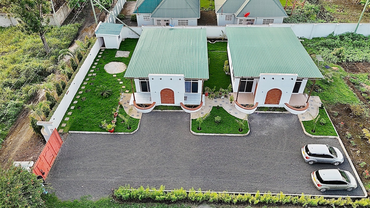 An aerial view showcases two adjacent buildings with green roofs, surrounded by well-maintained gardens. A gravel pathway leads to each entrance, while a circular stone feature is located in the nearby grassy area. Cars are parked along the driveway, enhancing the serene landscape.