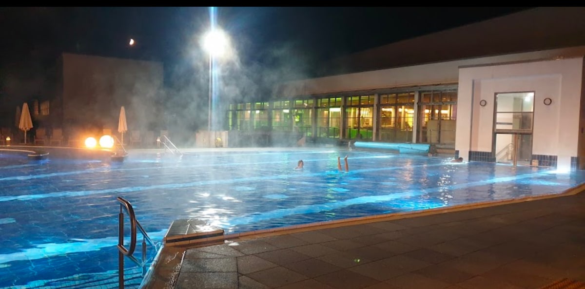 A large outdoor pool is illuminated at night, with steam rising from the water's surface. Several figures can be seen enjoying the warm water, while lounge chairs are arranged around the pool's perimeter. Bright lights from the surrounding building create a welcoming ambiance.