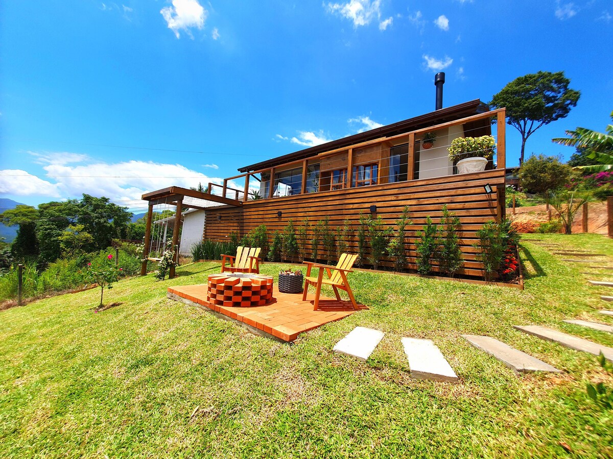 The exterior view showcases a modern cabin with large windows, set against a backdrop of lush hills. A wooden deck with seating and a fire pit invites outdoor relaxation, surrounded by manicured lawn and flowering plants, enhancing the serene landscape.