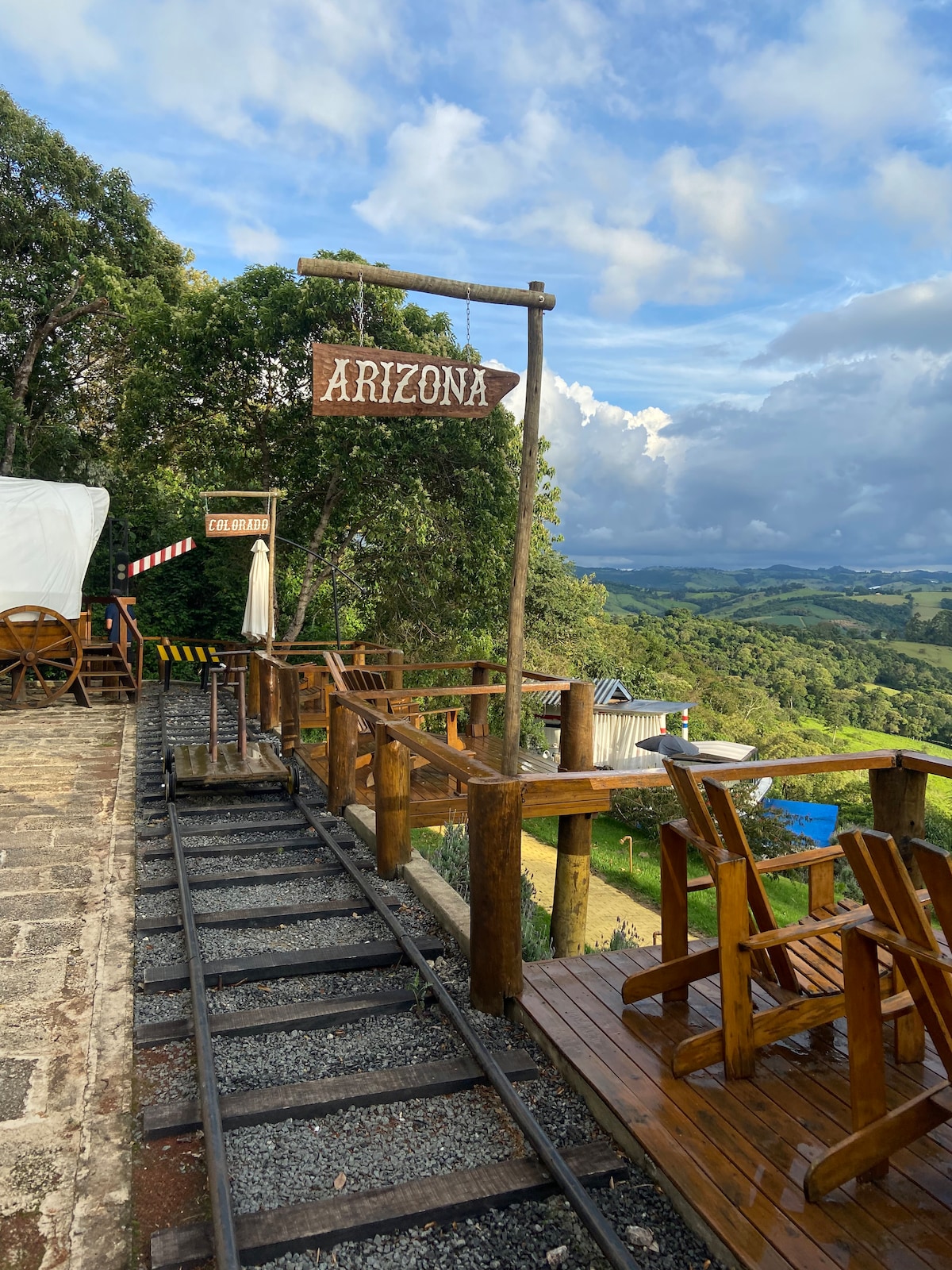 A wooden deck extends along a train track, with a sign labeled 'Arizona' positioned prominently. The surrounding scenery features lush green hills under a partly cloudy sky, complemented by wooden chairs for seating. The layout showcases a blend of rustic charm and natural beauty.