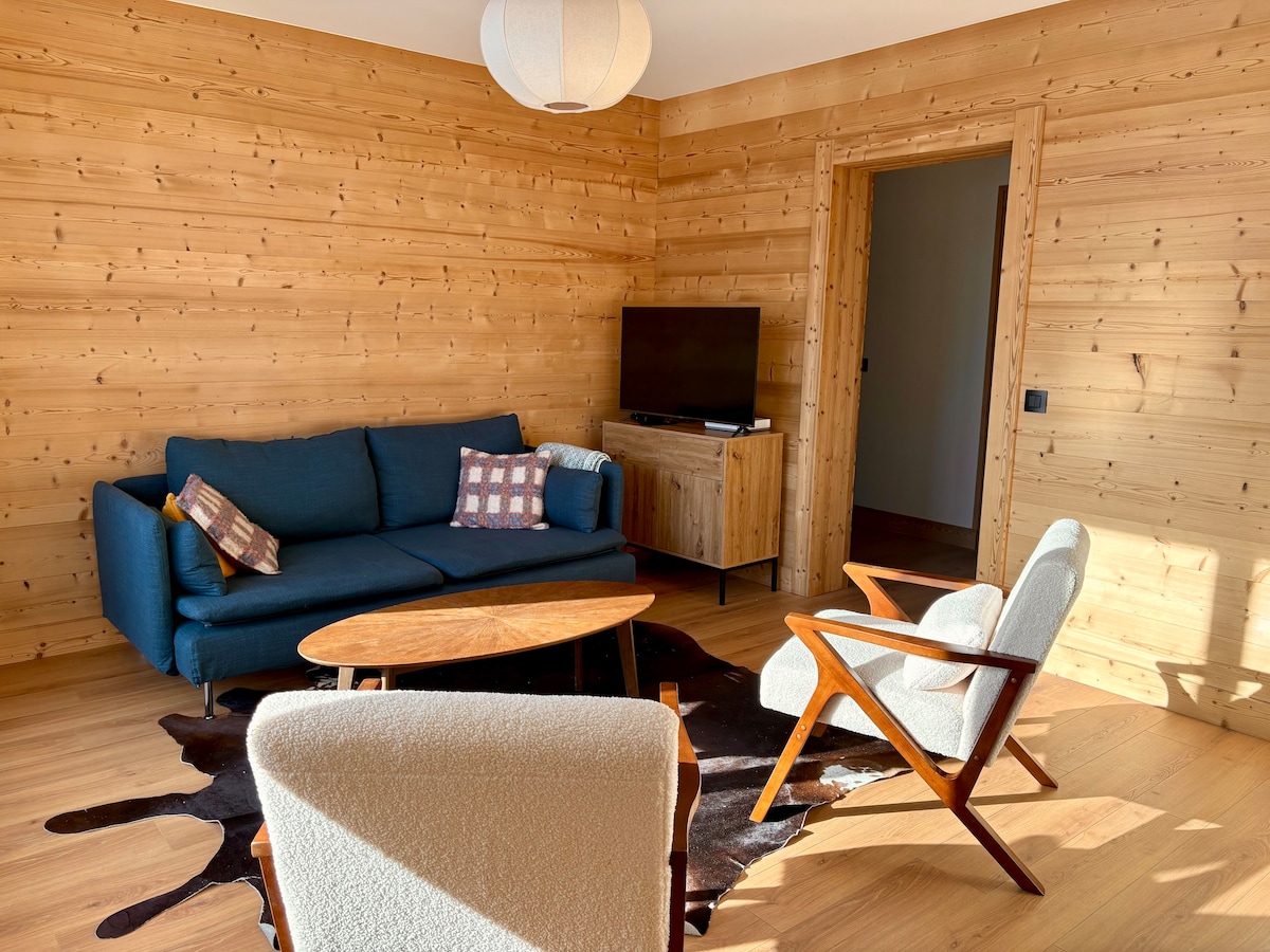 A spacious living area features wooden walls and a warm light from a hanging lamp. A blue sofa is positioned next to a wooden coffee table, with two mid-century style chairs nearby. A television is mounted against the wall, offering a cozy atmosphere.