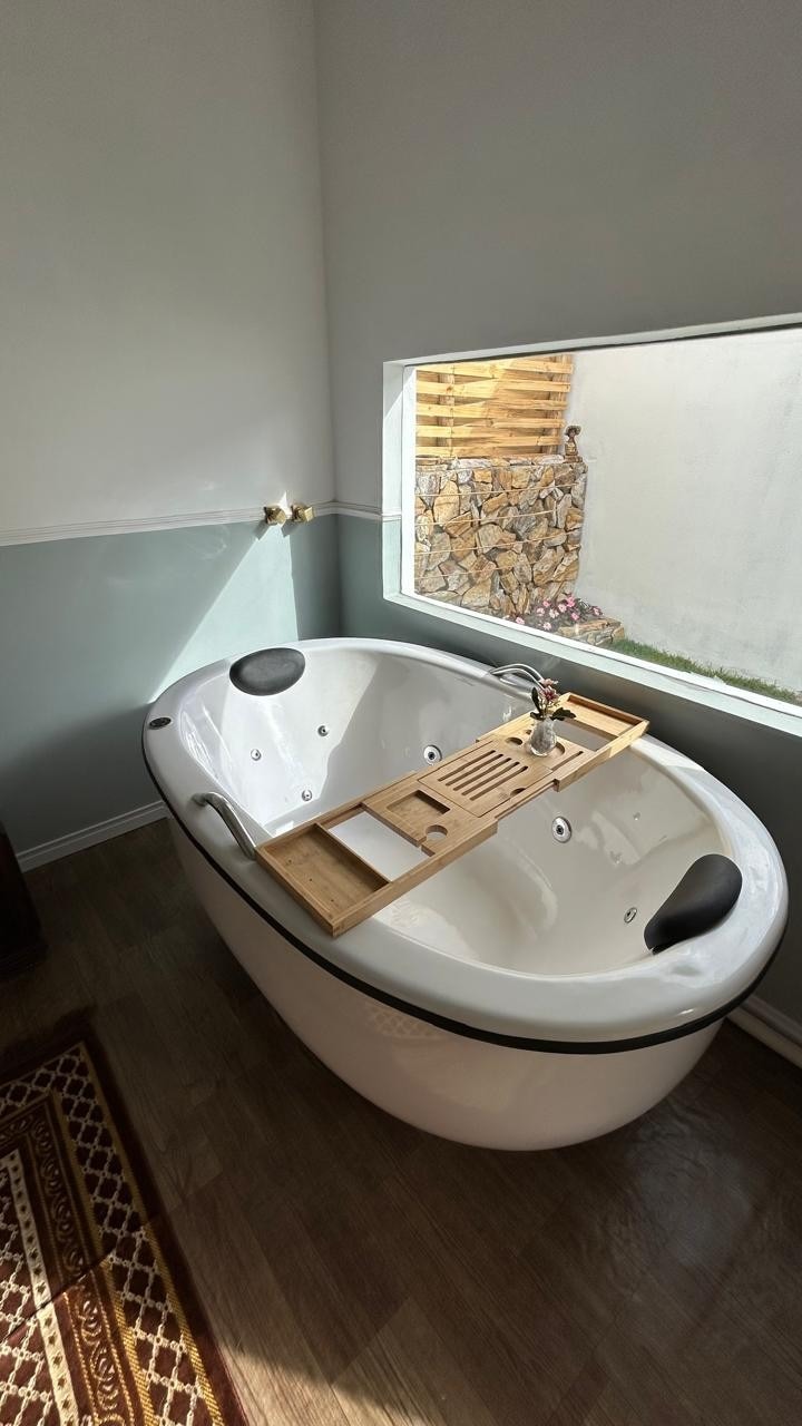 A modern, oval bathtub is positioned near a large window, inviting natural light into the space. A wooden bath caddy rests across the tub's edge, providing a stable surface for items. The surrounding walls are painted in soft tones, enhancing the serene atmosphere.