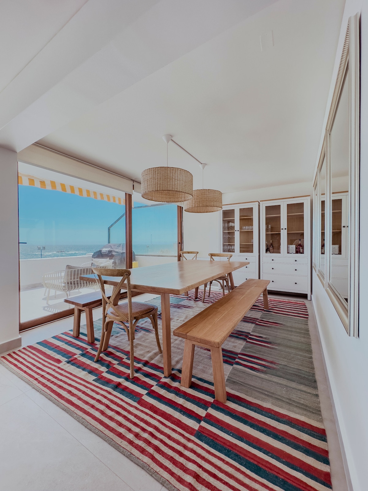 A dining area features a large wooden table surrounded by chairs, complemented by a long bench. Two woven pendant lights hang above, while a colorful striped rug lies beneath. Large windows offer views of the ocean, allowing natural light to illuminate the space.