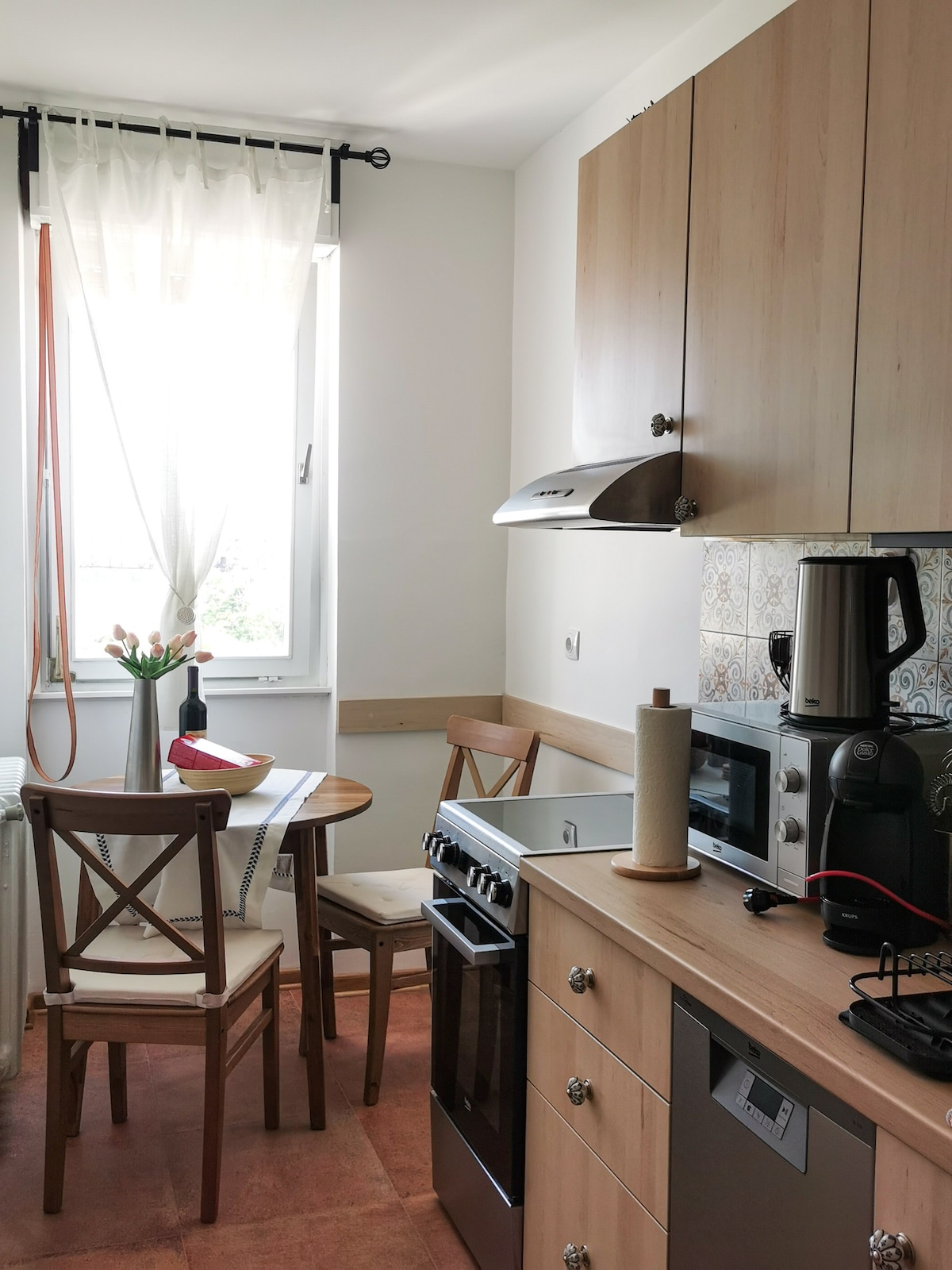 A compact kitchen features wooden cabinetry and modern appliances. A small round table is set for two with a fruit bowl. Natural light streams in through a window adorned with sheer curtains, highlighting the warm tones of the decor.