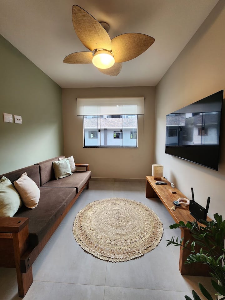Sala com sofa em suede, almofadas, tapete de palha,  aparador com decoração, abajur, TV 55", ventilador de teto