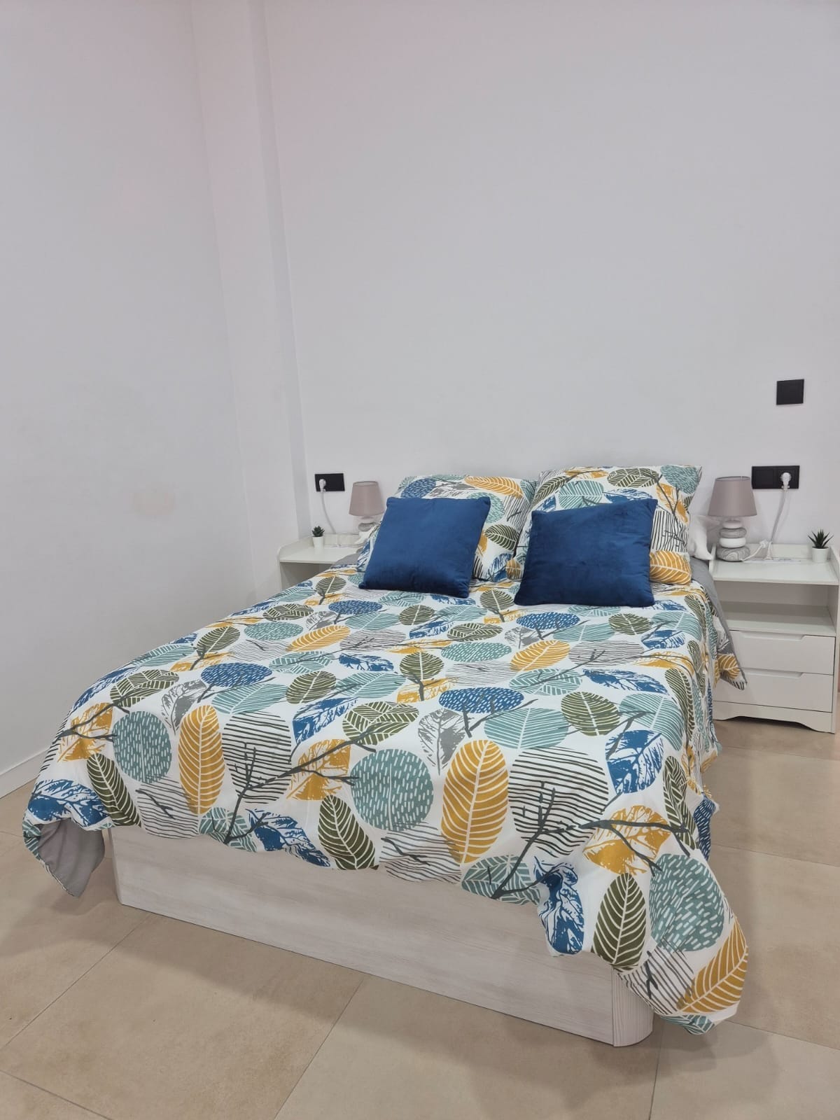 A bright sleeping area features a spacious bed with a colorful leaf-patterned bedspread. Two blue accent pillows are placed at the head of the bed. Simple nightstands are situated on either side, enhancing the room's minimalist design.