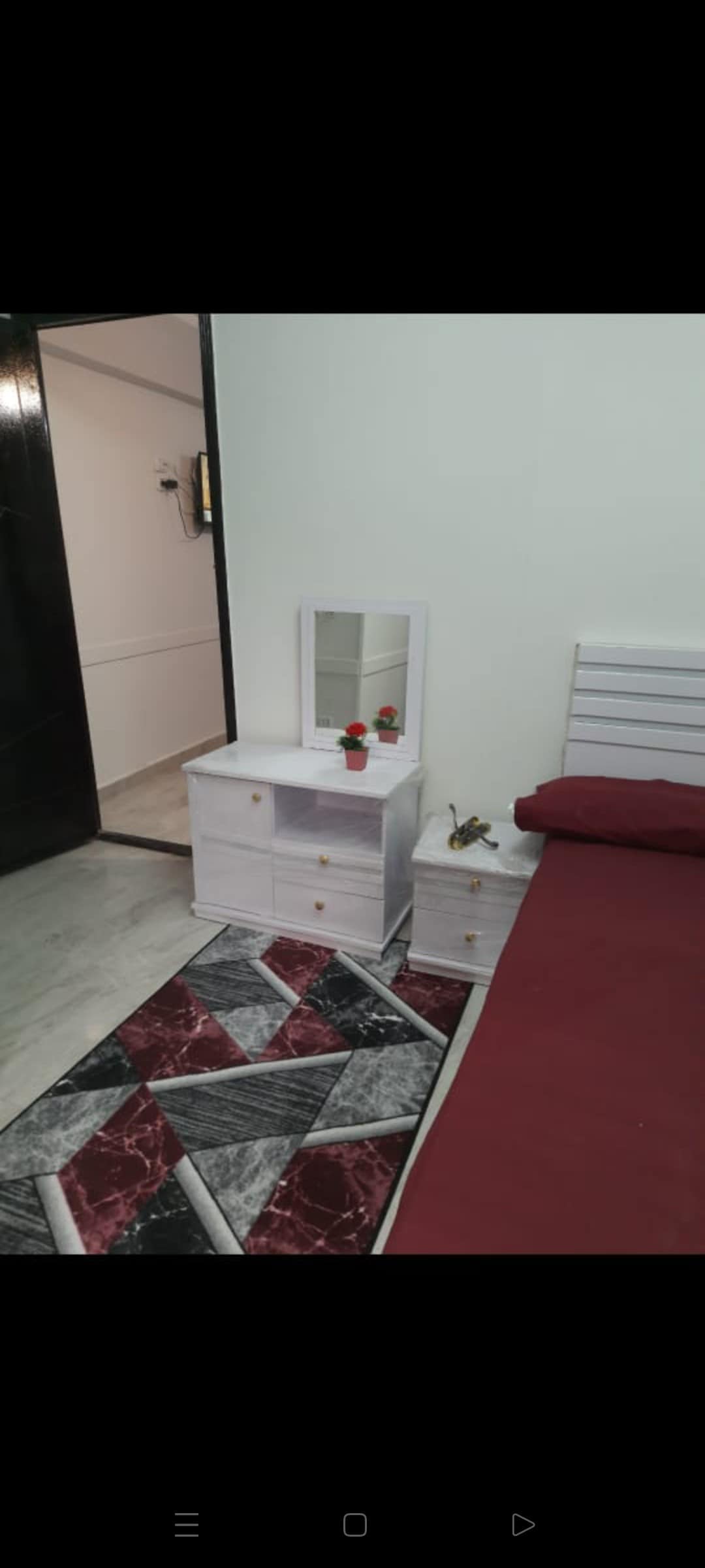 A simple bedroom setting is depicted with a spacious bed covered in a deep red blanket. A white nightstand with drawers is positioned beside the bed, featuring a small mirror and a decorative vase of flowers. A patterned rug adds texture to the floor.