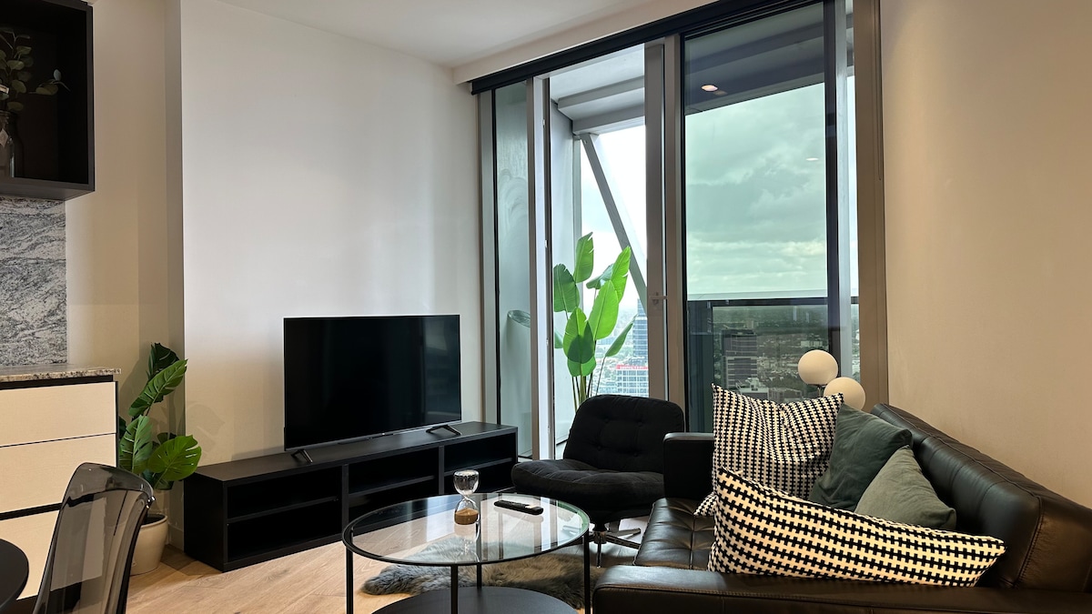 A modern living area is showcased with a comfortable leather sofa and sleek coffee table. A large television is mounted on a stylish cabinet, while floor-to-ceiling windows invite natural light and provide a view of the outdoors. Potted plants add a touch of greenery to the space.