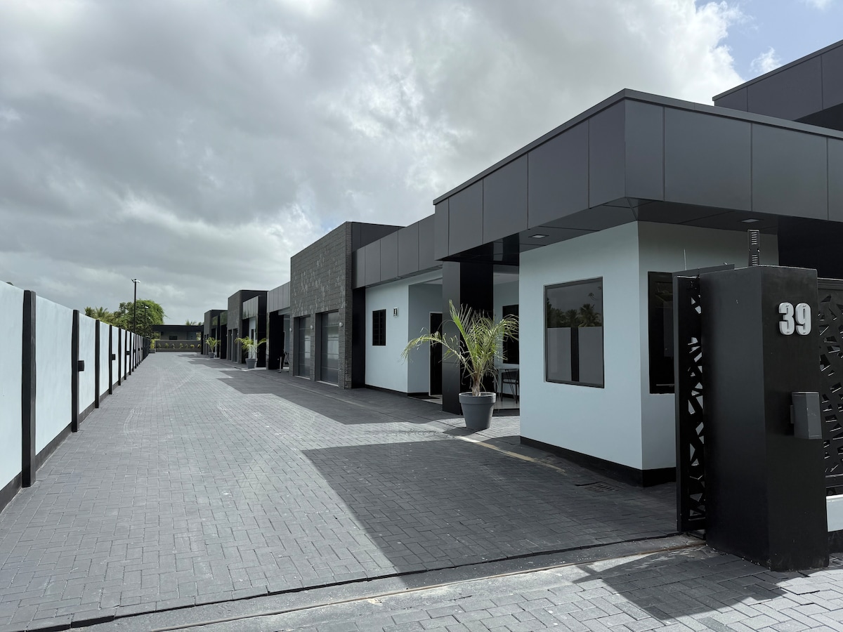 An exterior view of the property highlights a series of modern, one-story units arranged along a paved pathway. The minimalist architecture features dark accents and large windows. Potted plants add a touch of greenery, while the entrance gate is marked with the number 39.