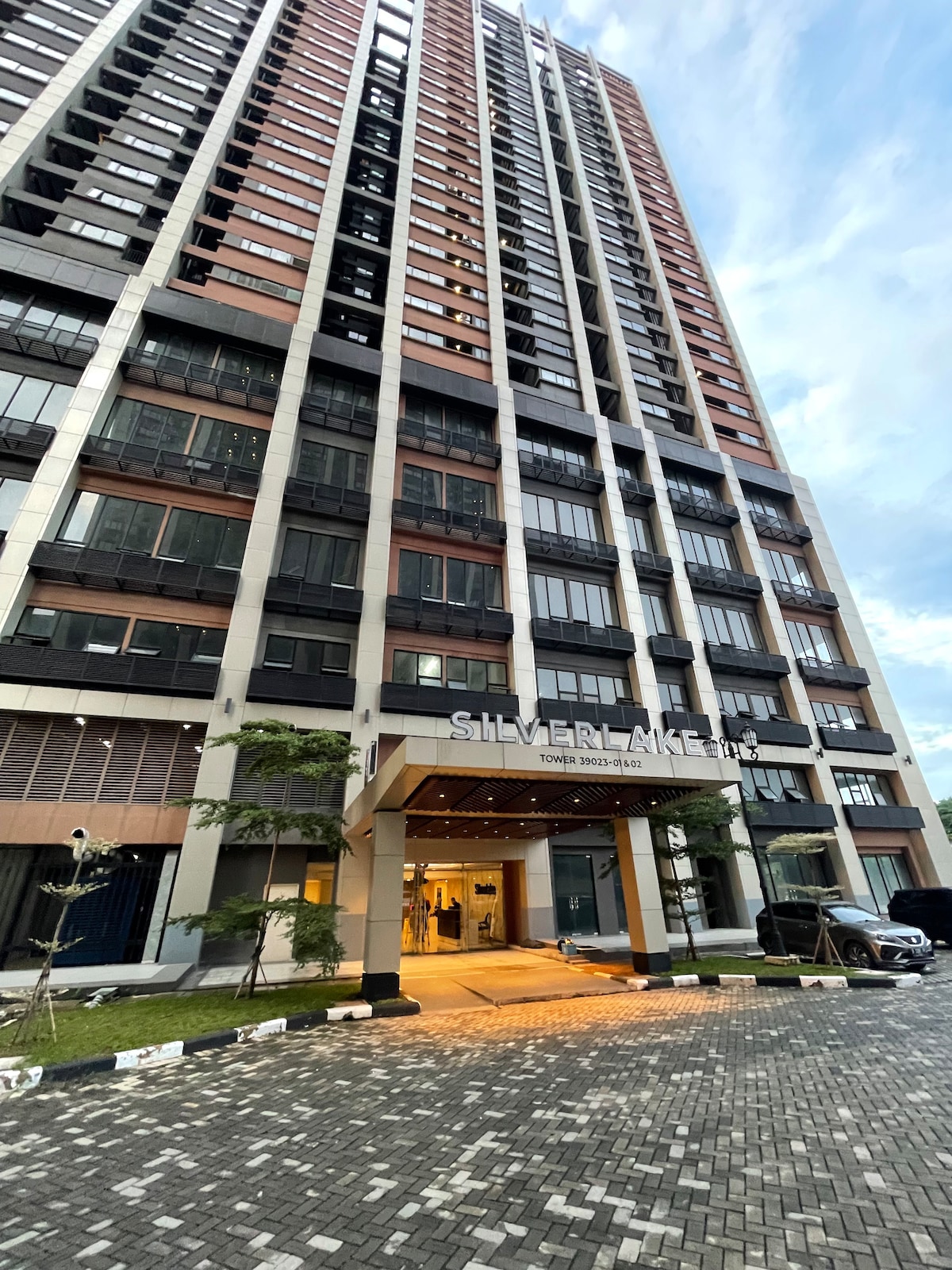 A modern high-rise building is presented, featuring a sleek exterior with a combination of glass and stone. The entrance showcases a welcoming foyer, and neatly landscaped pathways lead to the front, creating an accessible entryway for guests.