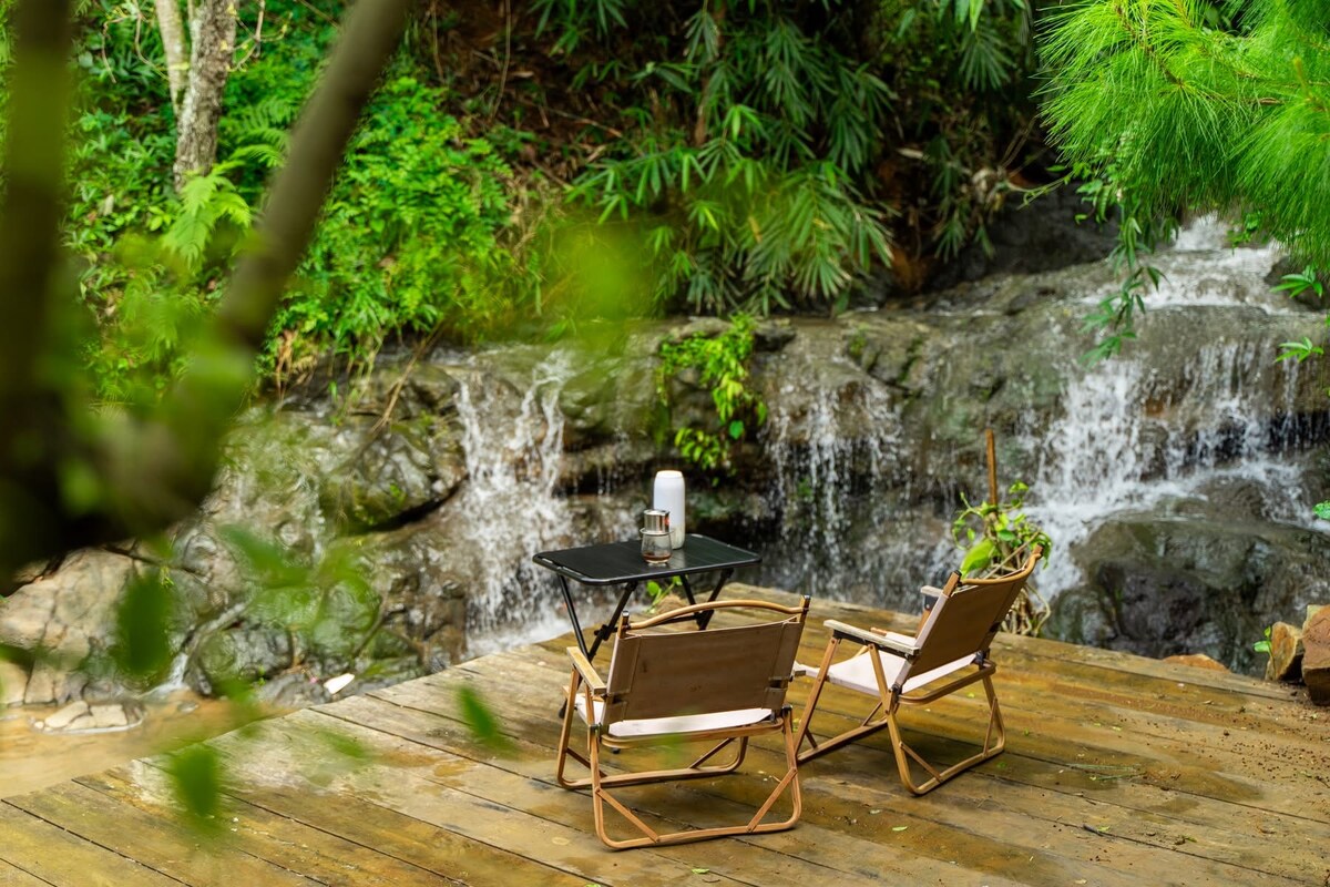 A wooden deck is positioned by a gently flowing stream, surrounded by lush greenery. Two folding chairs and a small table are set up, inviting relaxation. The soothing sound of the stream complements the tranquil natural setting.