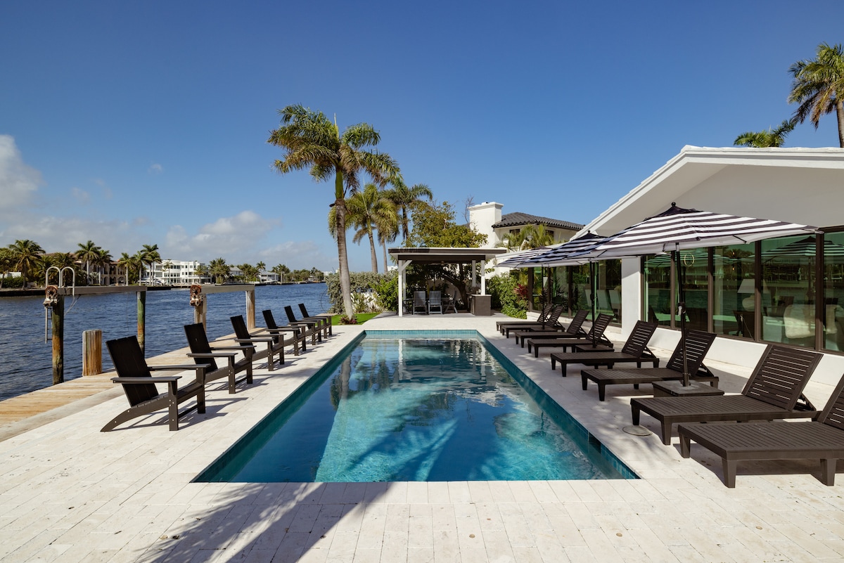 A heated pool area features sun loungers arranged along a stone deck, facing the Intracoastal waterway. Overhead, striped umbrellas provide shade, while palm trees enhance the tropical ambiance. The pool reflects clear blue skies and offers direct access to scenic waters.