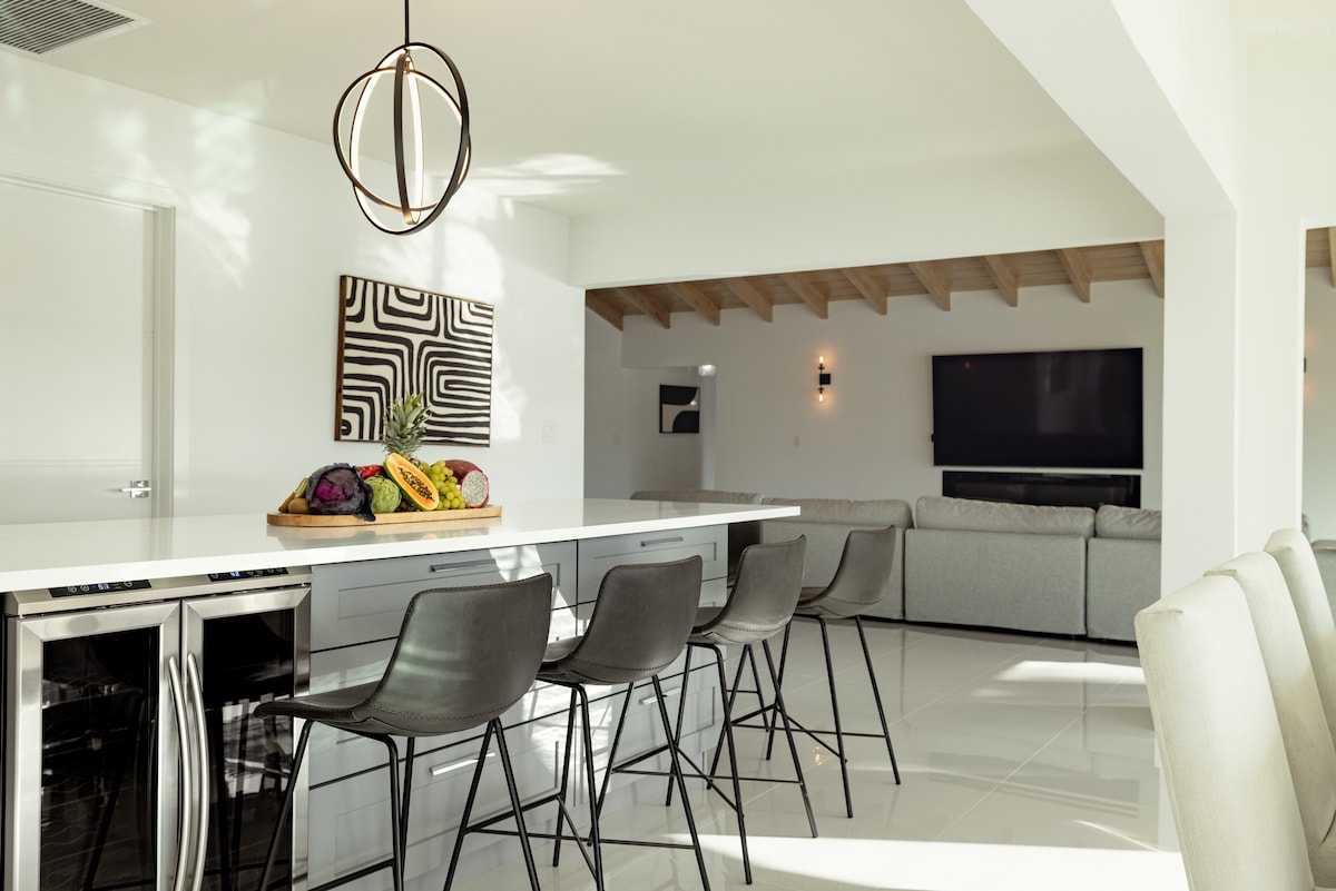 A modern kitchen area is highlighted by sleek countertops and a bar with high stools. A fruit centerpiece adds a touch of color. The open layout flows into a cozy living space, featuring a large sectional couch and a mounted television on the wall.