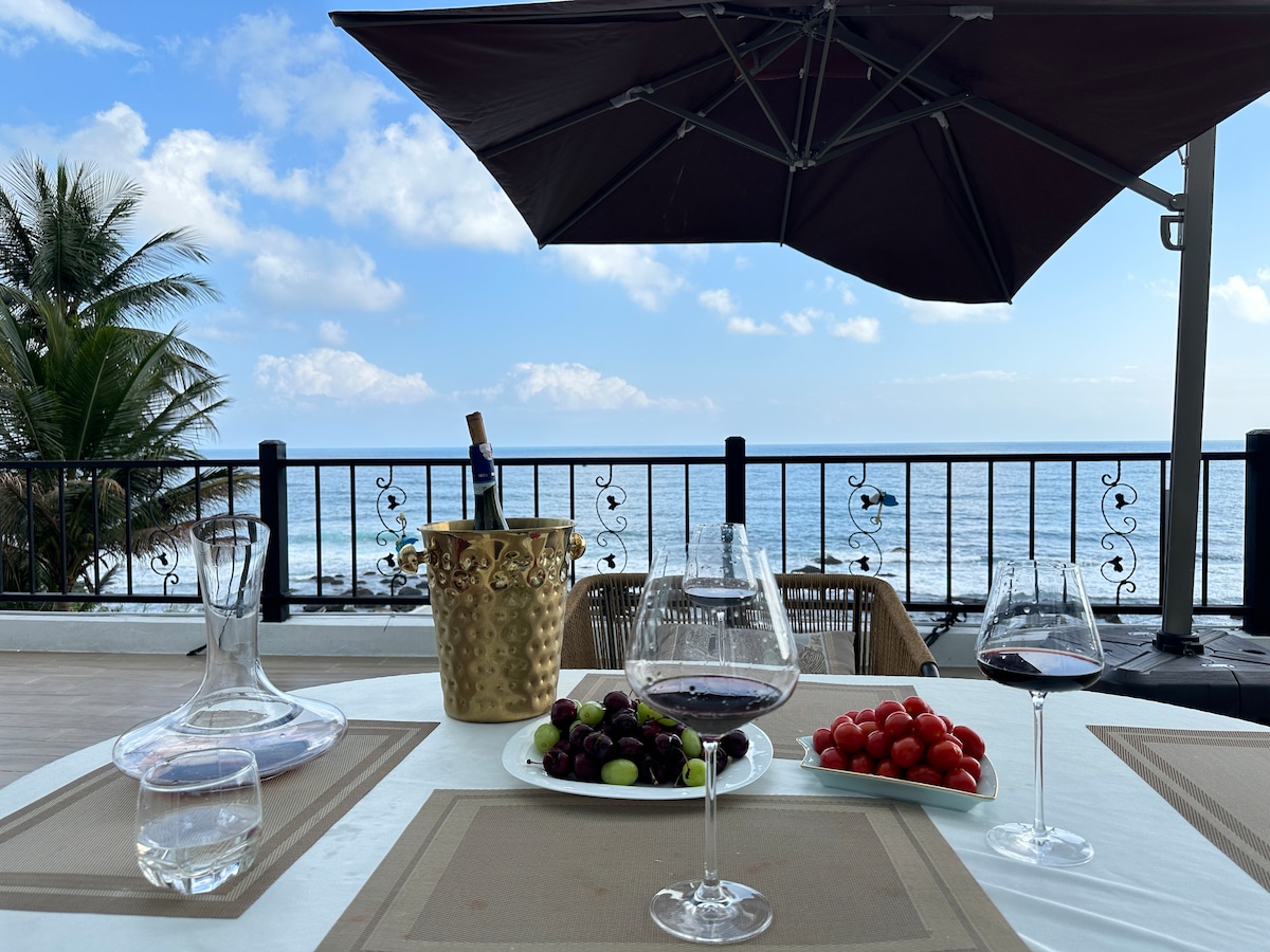 An outdoor dining table is set with glasses of red wine, a decanter, and a plate of fresh grapes and cherry tomatoes. A gold champagne bucket rests beside a backdrop of ocean waves and a clear sky, shaded by a large umbrella.