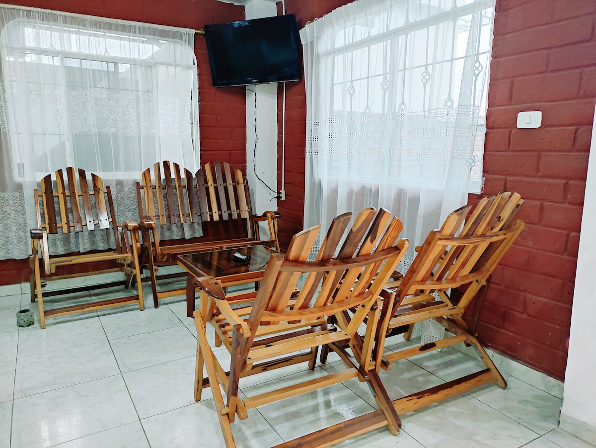 A cozy seating area features four wooden recliners arranged around a glass coffee table. A television is mounted on the wall, and natural light filters through sheer curtains covering a large window, creating a warm and inviting space.