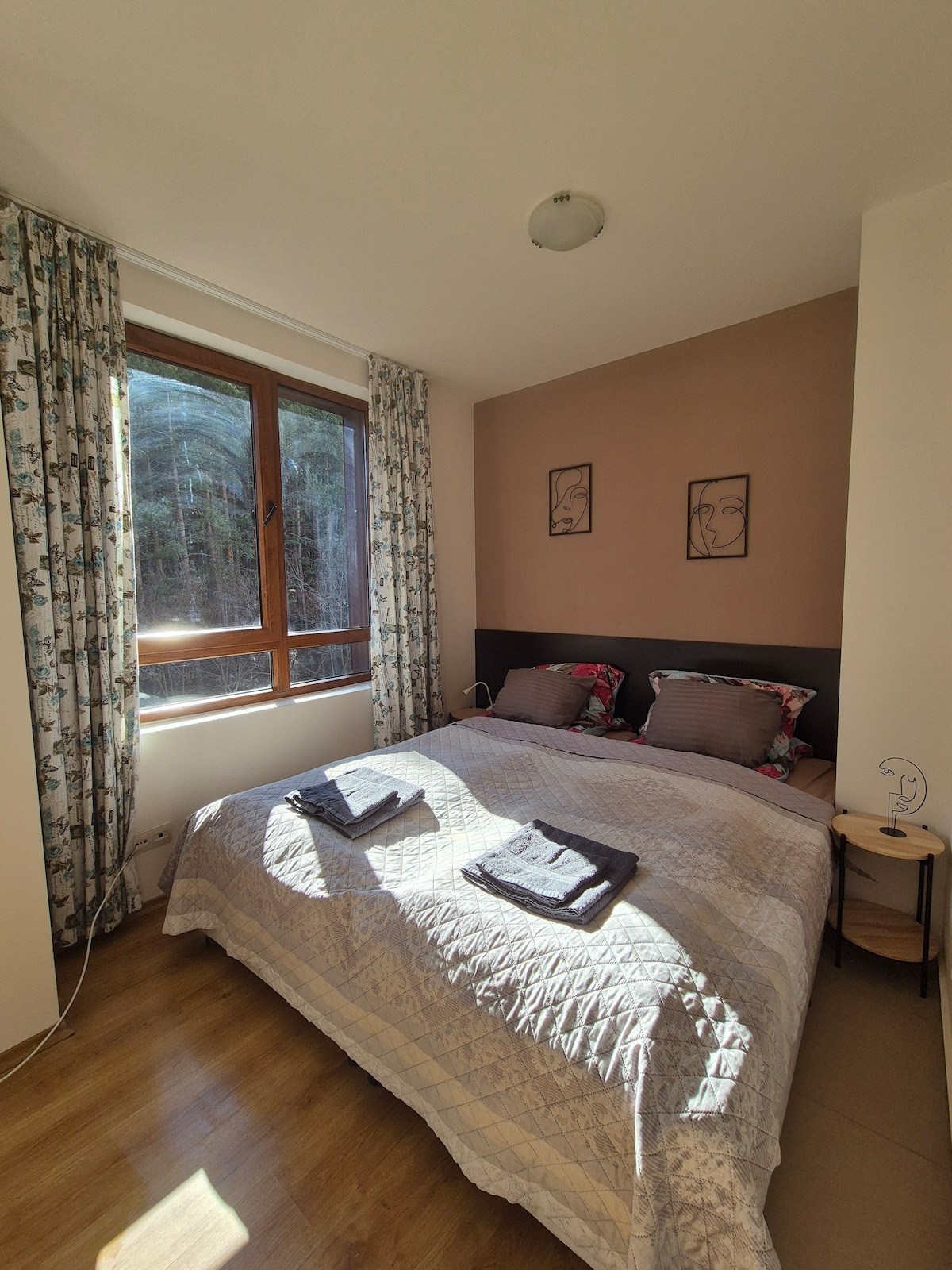 A well-lit bedroom is furnished with a spacious bed layered with soft linens and decorative pillows. Natural light enters through a large window adorned with patterned curtains. A bedside table holds a lamp, and fresh towels are neatly arranged on the bed.