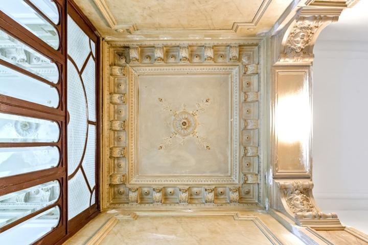 The image highlights an ornate ceiling featuring intricate moldings and decorative elements, illuminated by natural light. The wooden door at the edge provides a glimpse of the entryway, enhancing the historical charm of the heritage building.