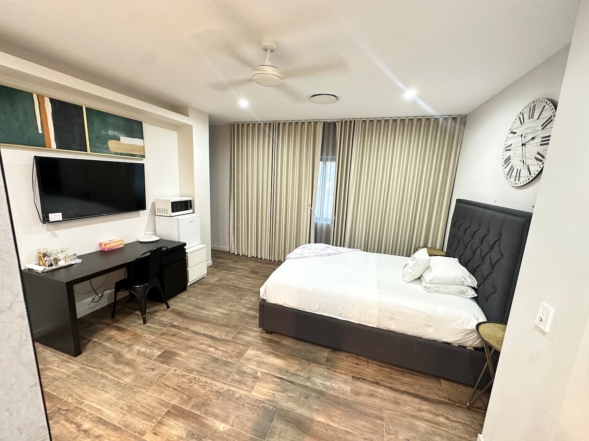 A modern room features a spacious bed with crisp white linens. A large wall-mounted television and a compact microwave are positioned on a sleek cabinet. A wooden desk and chair provide a workspace. Light flows through floor-length curtains that frame the window.