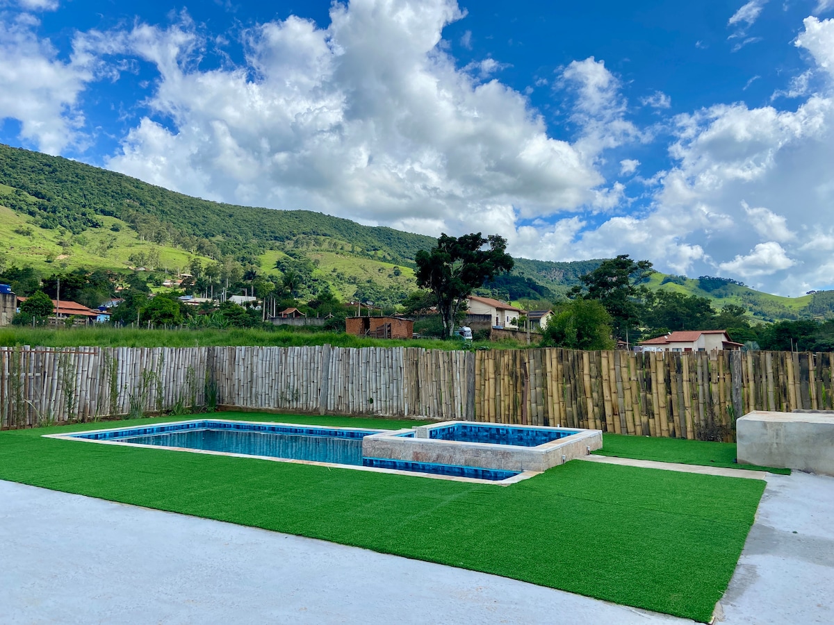 A spacious outdoor area features a well-maintained lawn and a swimming pool, complemented by a hot tub. A bamboo fence provides privacy while the scenic mountains create a serene backdrop. Bright blue skies and decorative clouds enhance the tranquil atmosphere.