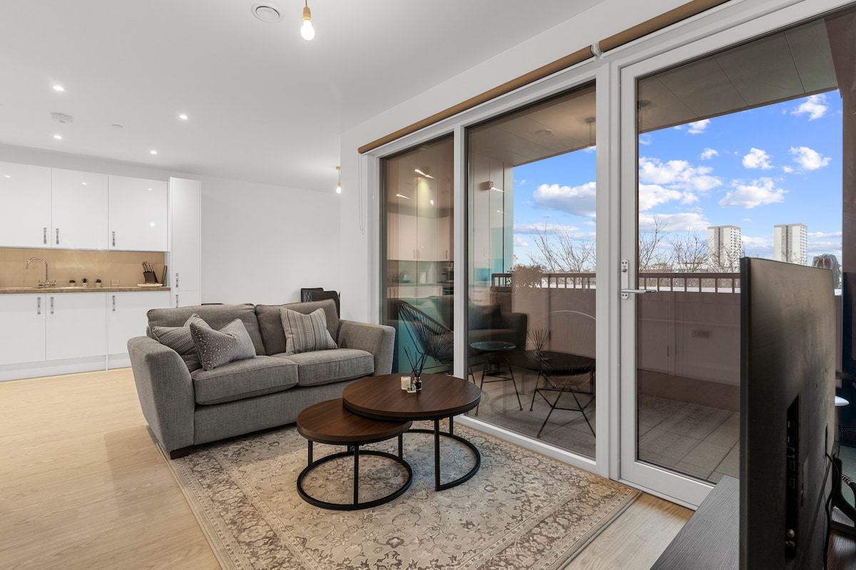 A modern living area is visible, featuring a comfortable grey sofa and two circular coffee tables. Large glass doors open to a balcony, allowing natural light to fill the space. A kitchenette with white cabinetry is located in the background, enhancing the contemporary design.
