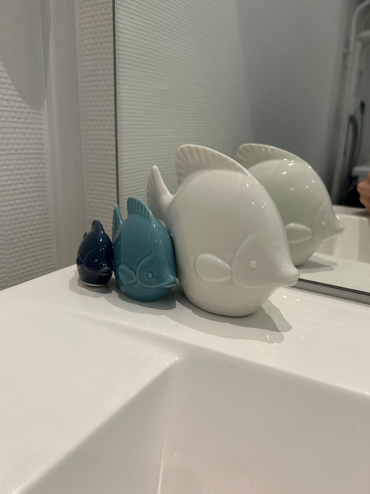 A collection of ceramic fish decorations rests on a white sink. The fish are arranged in varying sizes, with a large white fish in the back and two smaller fish in shades of blue in front. A bathroom mirror is partially visible in the background.