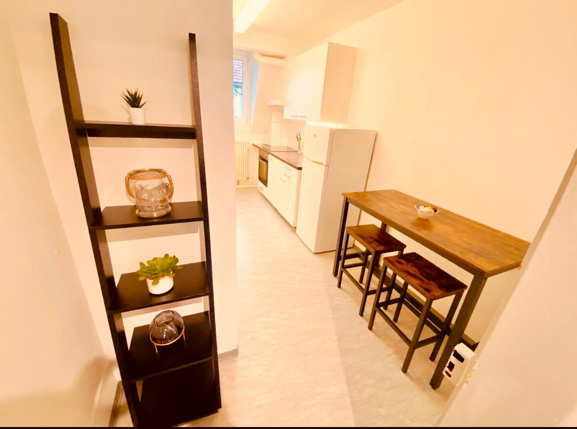 A modern kitchen area is presented with a sleek white refrigerator and stove. A wooden table with three high stools is positioned near a shelf displaying decorative items and plants. Natural light enhances the bright, neutral-colored walls, creating an inviting, functional space.